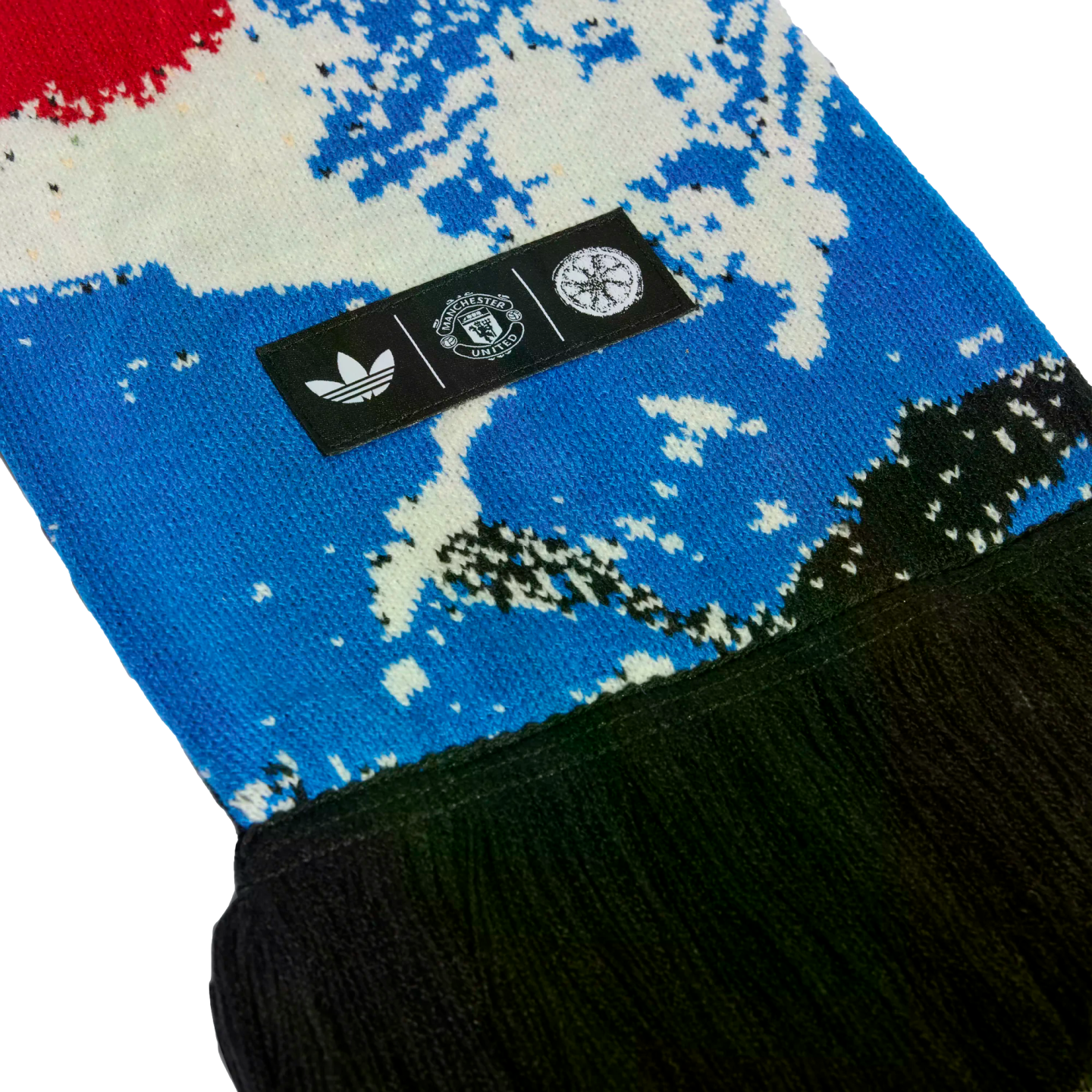 Close-up of a blue and white patterned knit scarf with black fringe. A black tag displays the white Adidas trefoil, Manchester United crest, and a circular logo.