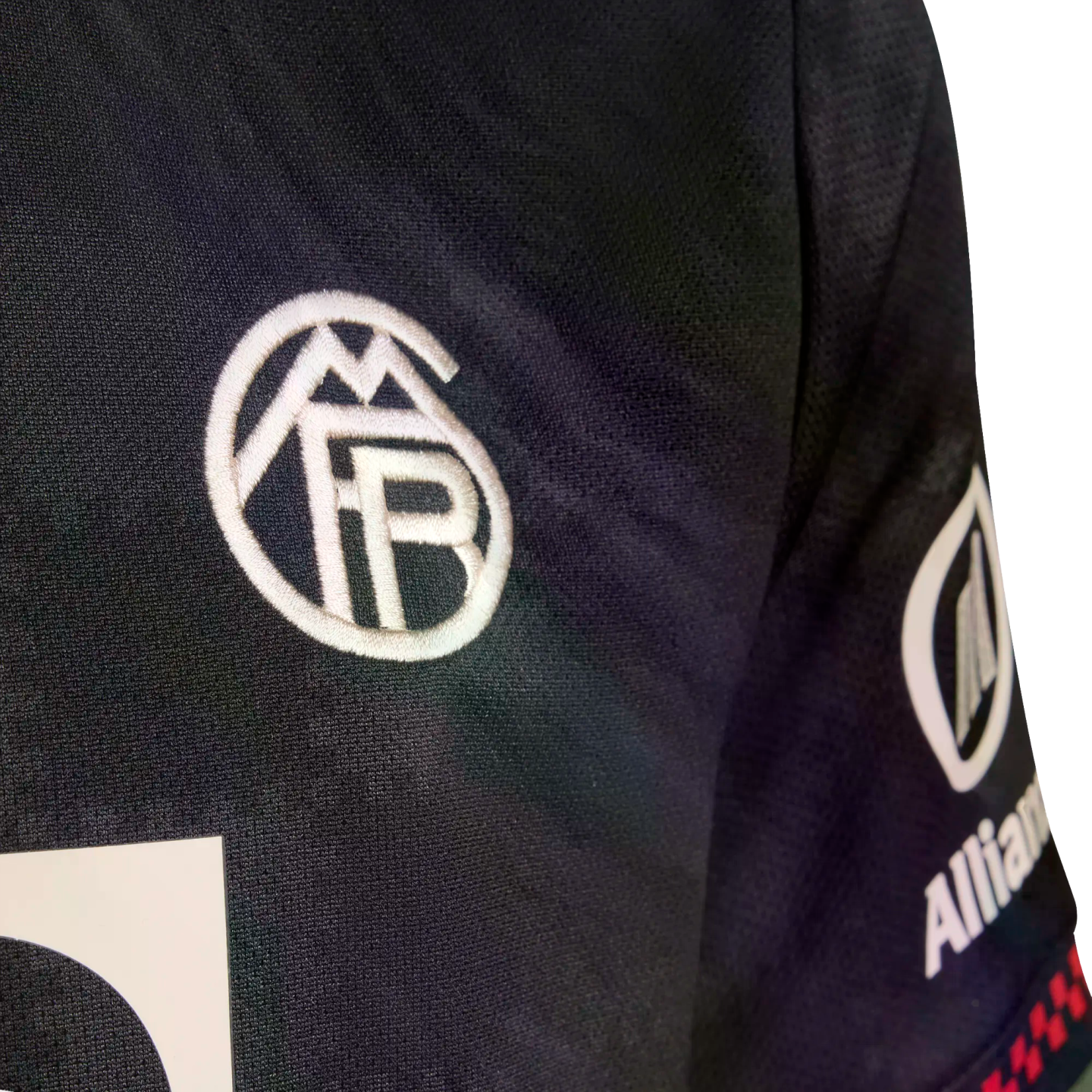 Close-up of a dark textured athletic jersey with a white circular embroidered logo on the chest and an Allianz sponsor logo on the sleeve.
