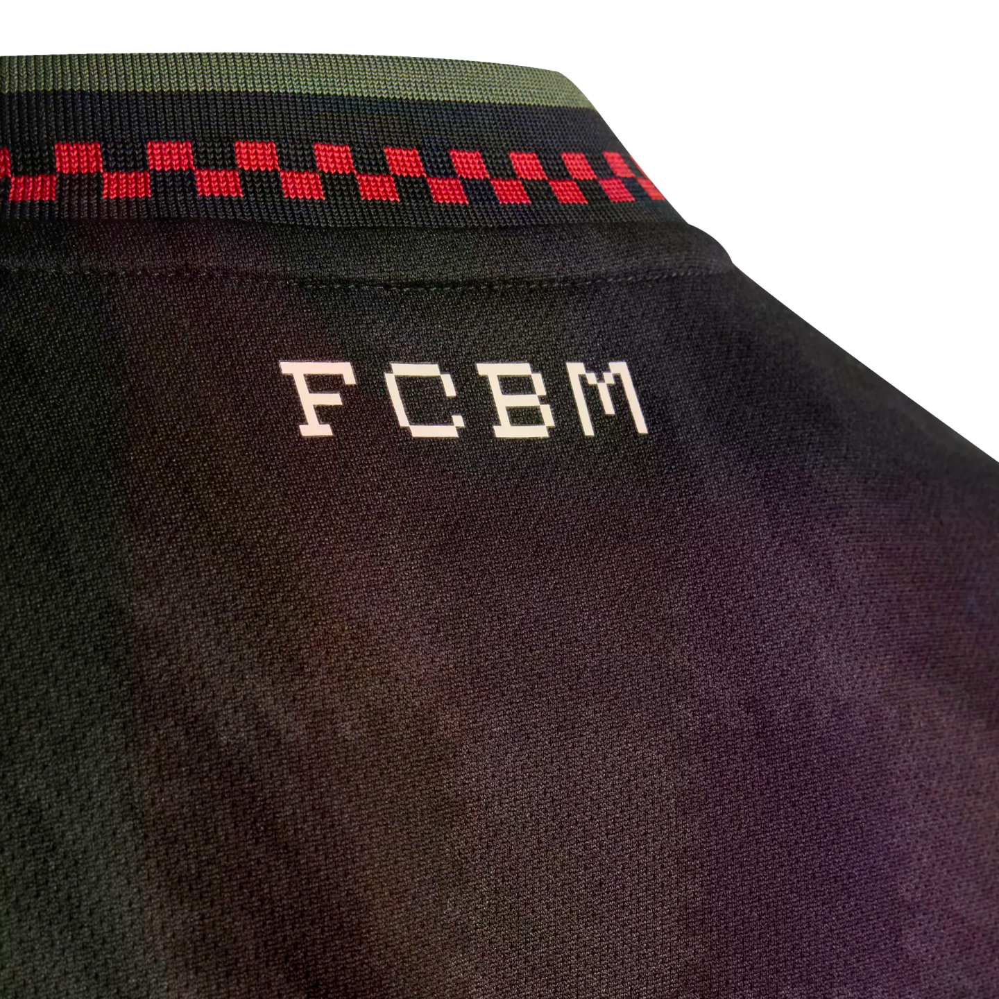 Close-up of a black jersey's back neck, showing white blocky "FCBM" text below a collar with green, navy, and red checkered patterns.