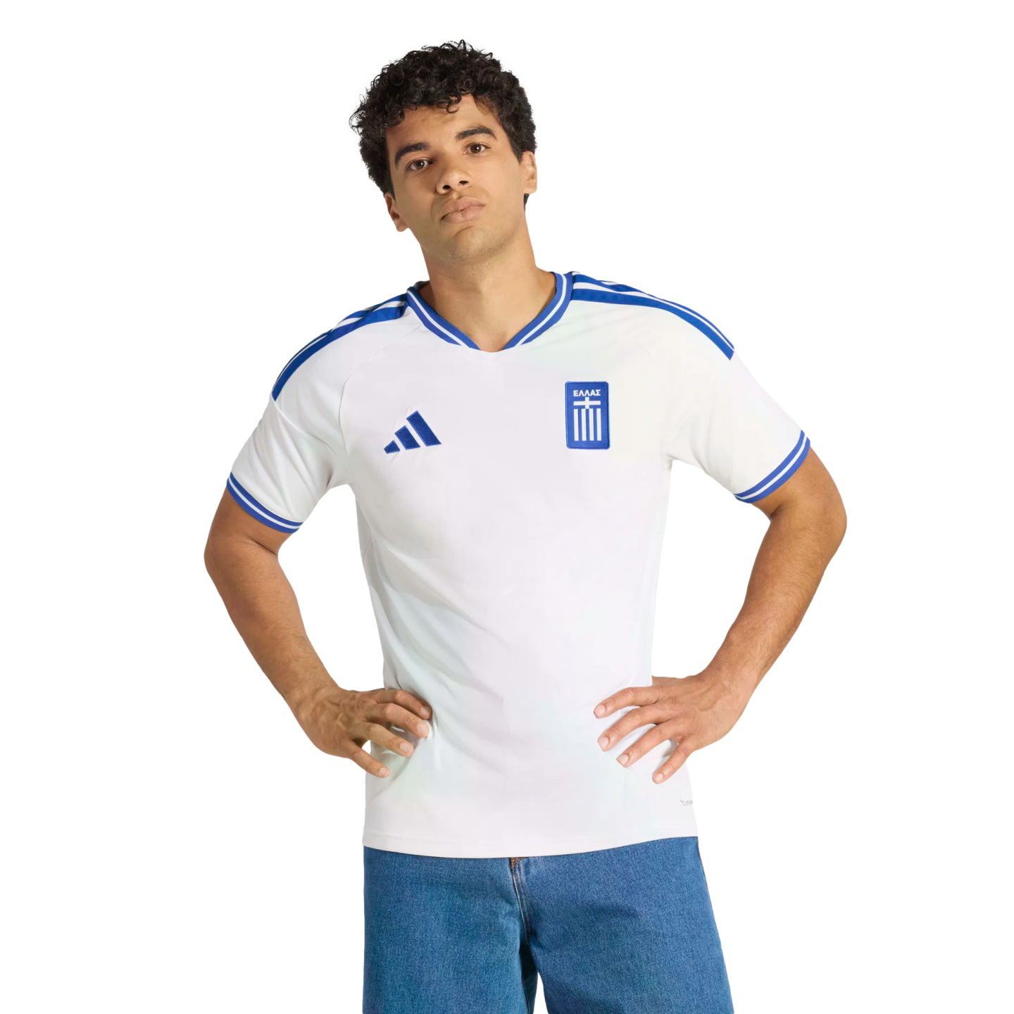 A man in a white Greece national team soccer jersey with blue trim stands with hands on his hips against a black background.