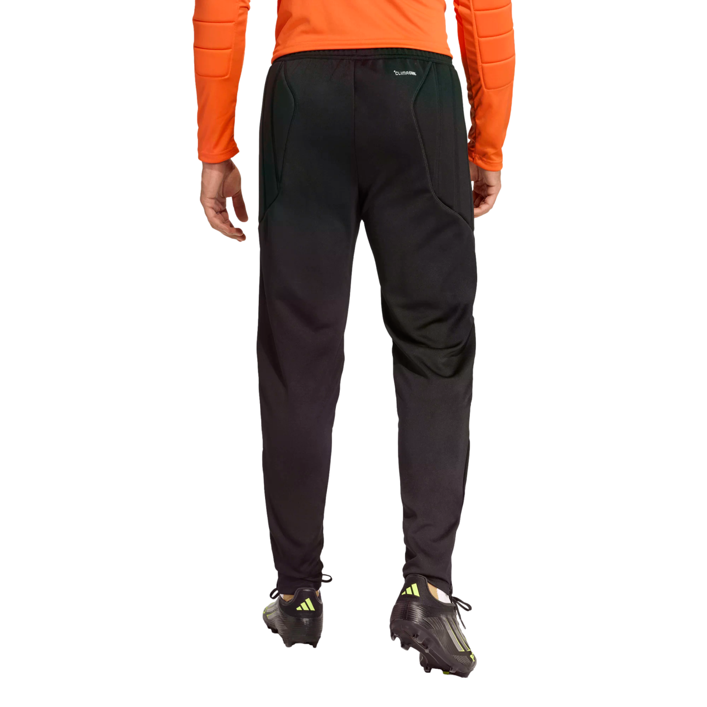 Rear view of a person wearing black Climacool athletic pants and soccer cleats, wearing an orange long-sleeved top against a black background.