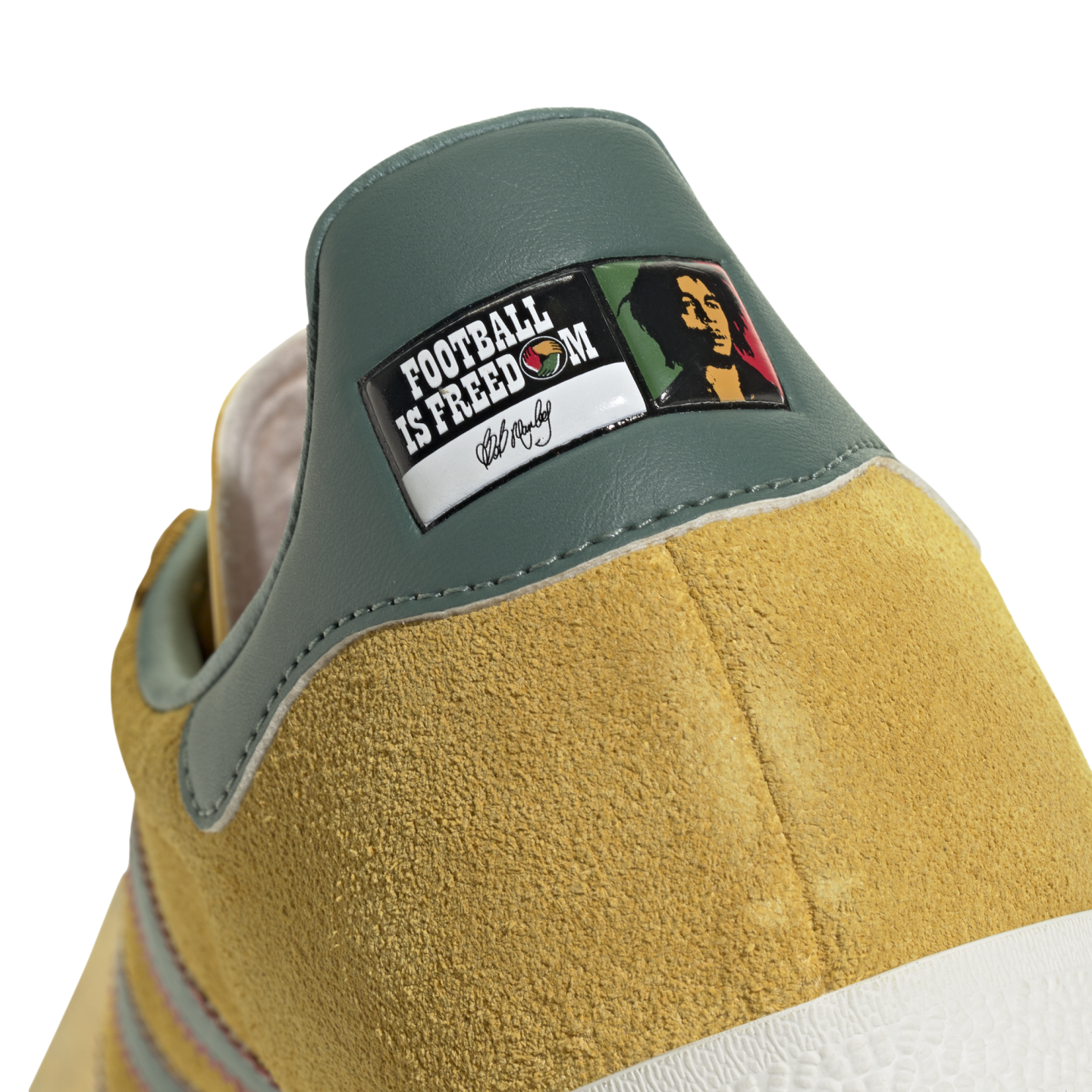 Close-up of a yellow suede shoe's green heel tab with a graphic patch reading "FOOTBALL IS FREEDOM," featuring Bob Marley’s portrait and signature.