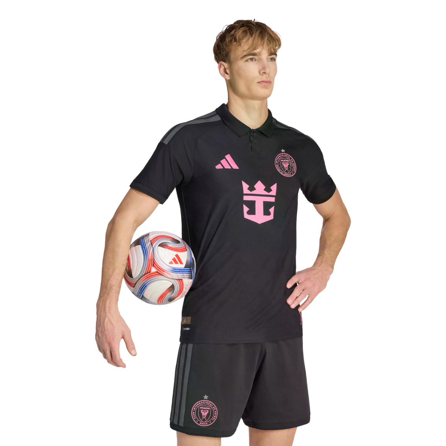 A male model wearing a black Inter Miami CF soccer kit with pink accents, holding a soccer ball under his arm against a plain black background.