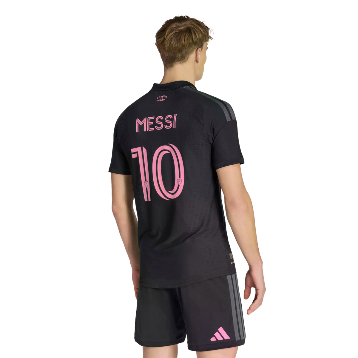 A person from behind wearing a black soccer jersey with the name "Messi" and the number "10" in pink on the back. He also wears matching black shorts with a pink Adidas logo on the leg, all set against a solid black background.