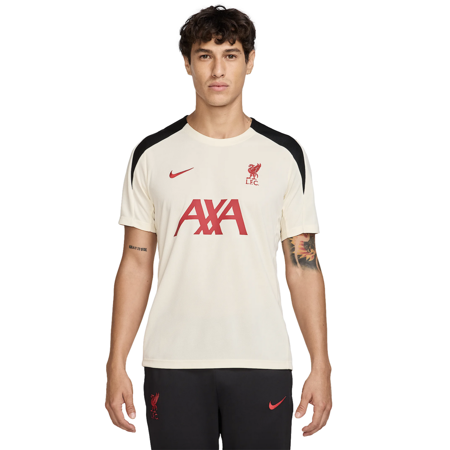 A male model wears an off-white Liverpool FC training jersey with red logos and black shoulder panels, paired with black Nike athletic pants.