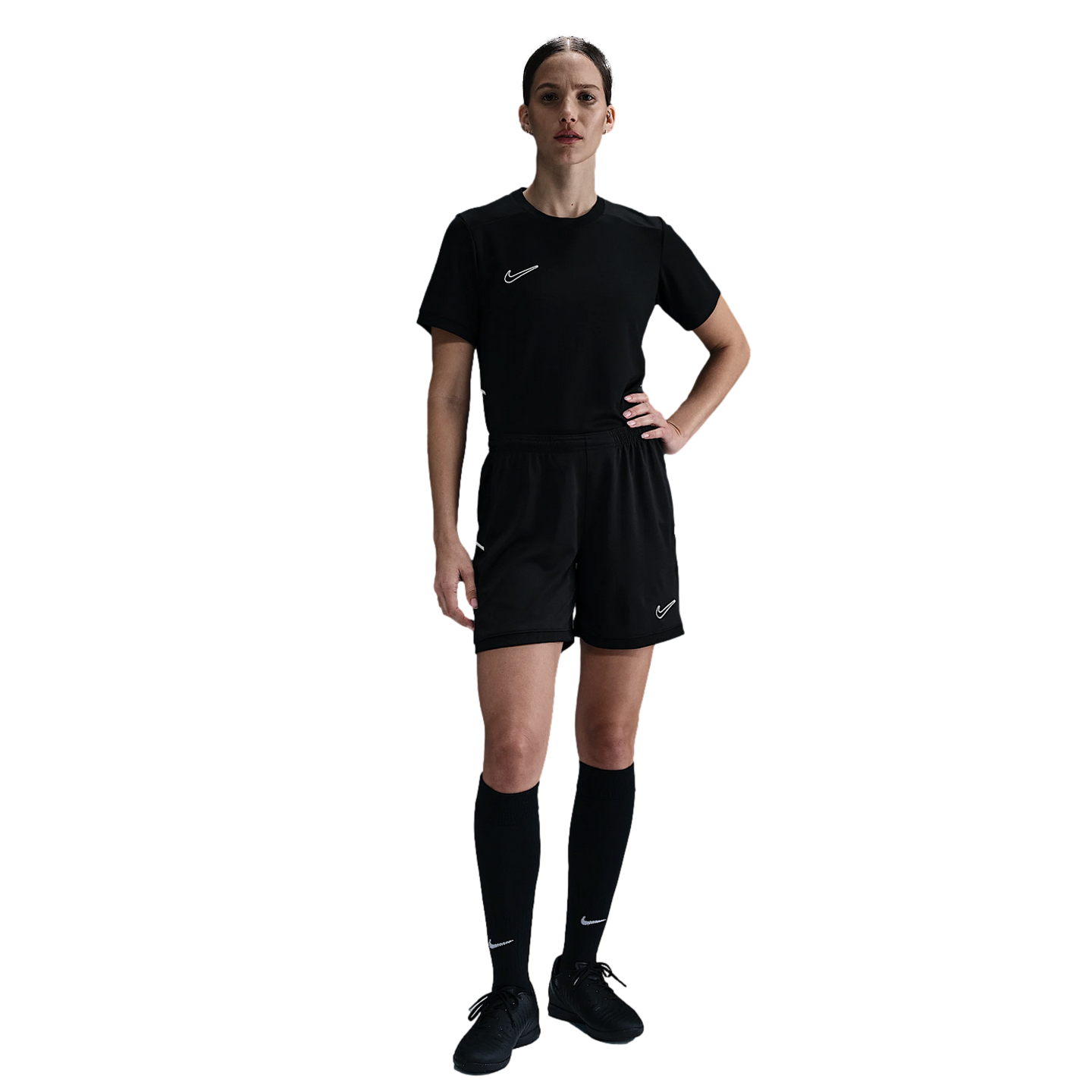 A woman in an all-black Nike soccer kit, including a jersey, shorts, knee-high socks, and cleats, stands against a plain white background with one hand on her hip.