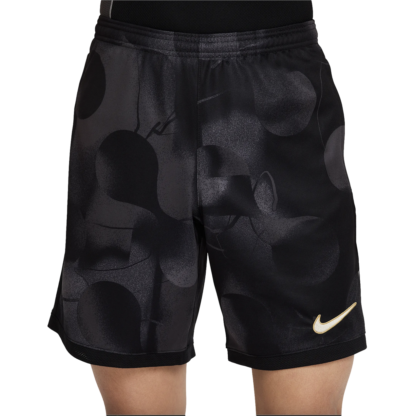 A person wearing black Nike athletic shorts with a dark grey abstract pattern and a gold swoosh logo on the leg.