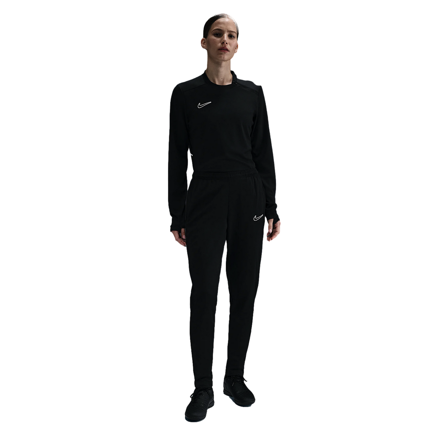 A woman standing in an all-black Nike tracksuit featuring a long-sleeve top and matching joggers, each with a small white swoosh logo, against a plain white background.