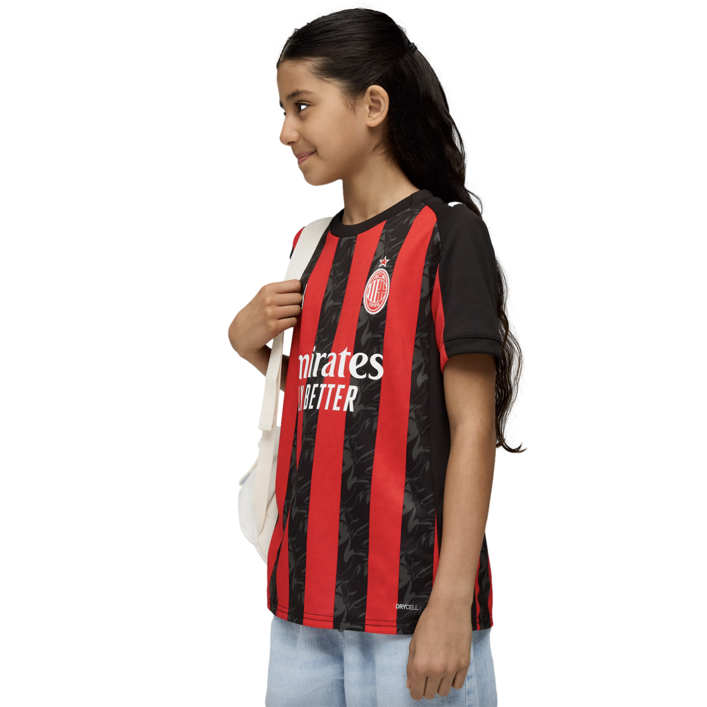 A young girl in a red and black AC Milan soccer jersey and light jeans smiles while carrying a white backpack against a white background.