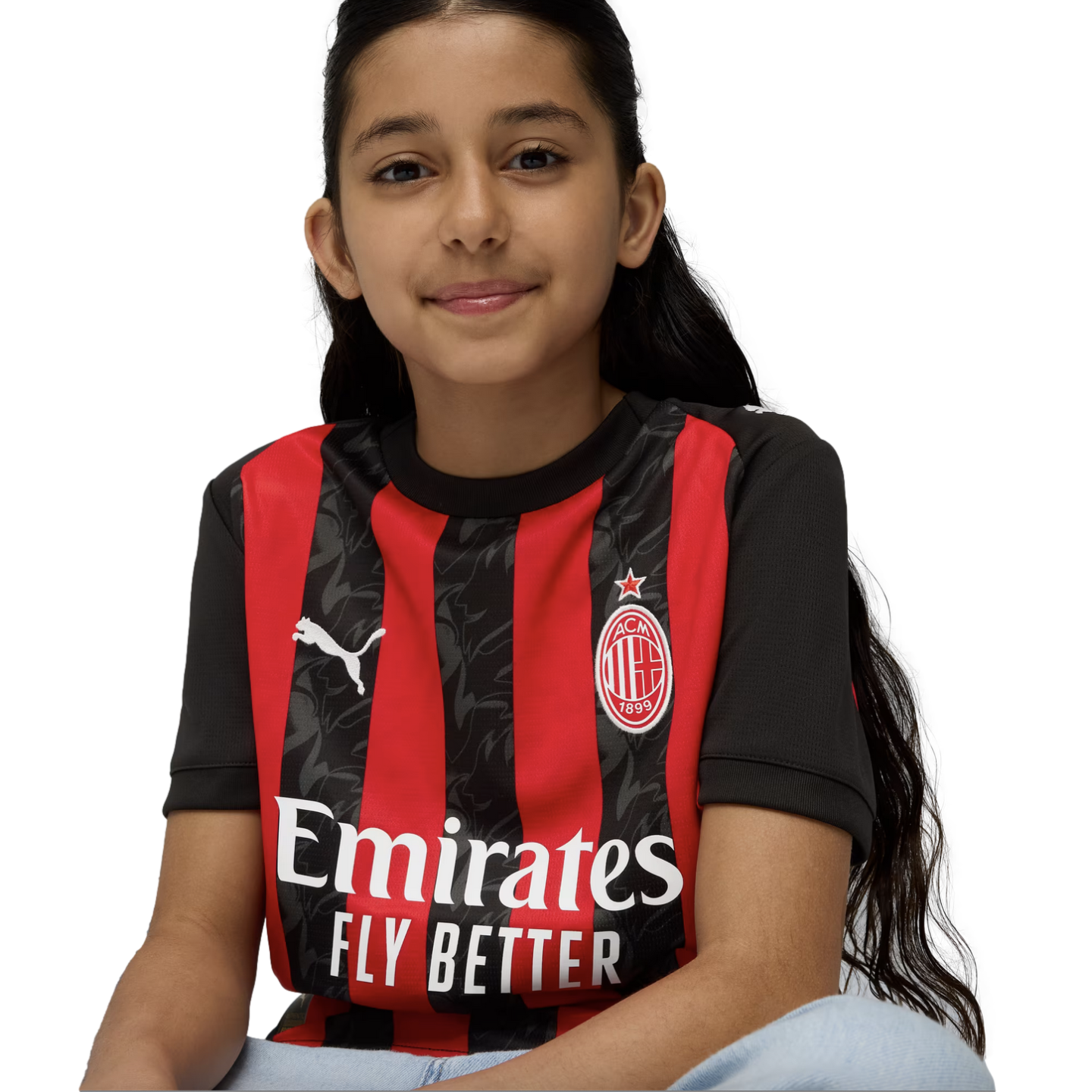 A smiling young girl with long dark hair sits against a white background wearing a red and black striped AC Milan soccer jersey.