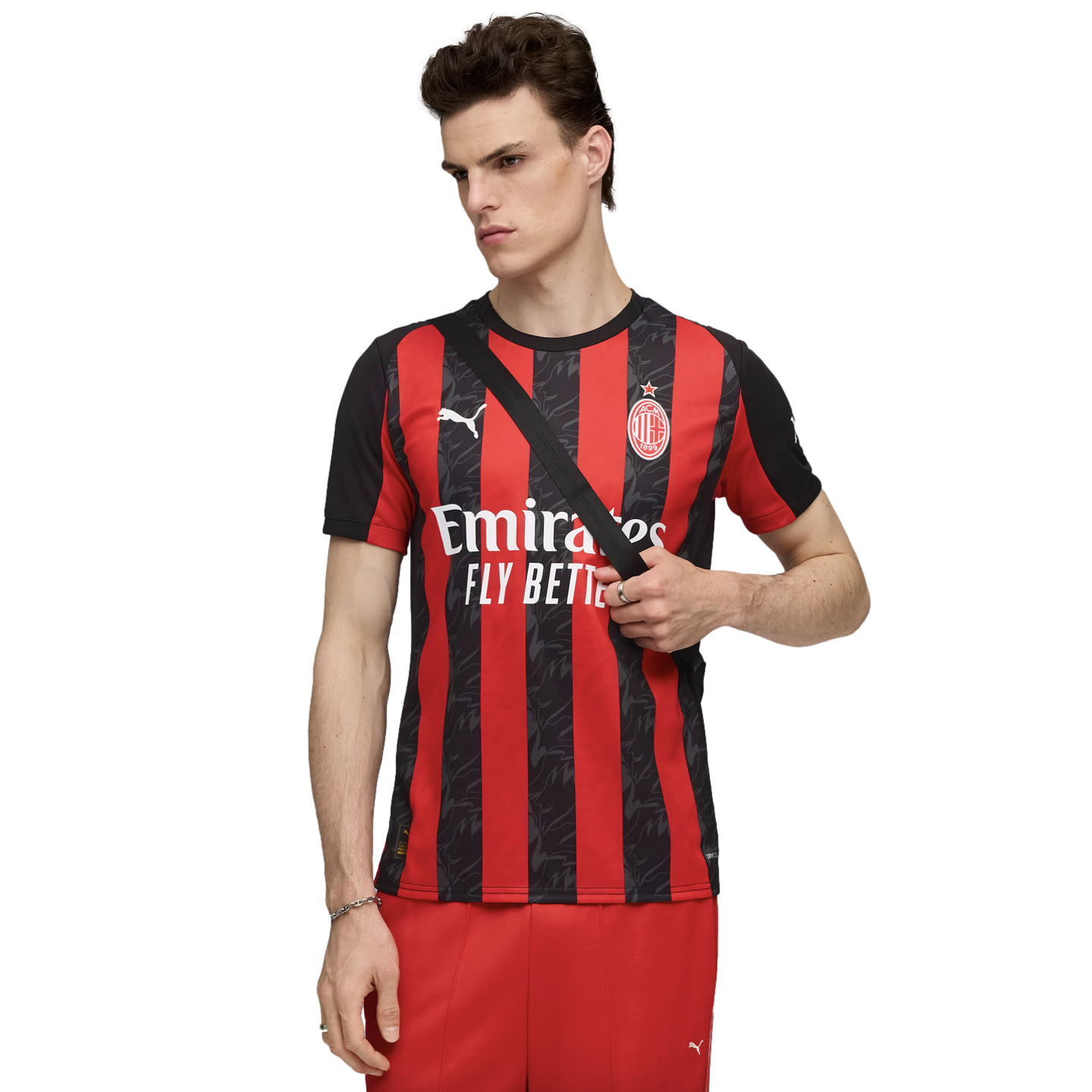 A male model wearing an AC Milan red and black striped home jersey with the Emirates sponsor logo, paired with red shorts and a black cross-body bag.