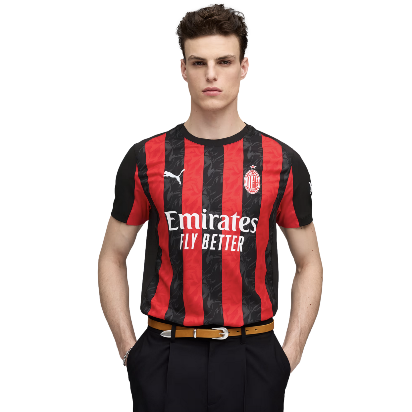 Young man wearing a red and black striped AC Milan soccer jersey, black trousers, and a tan belt, posing against a white background.