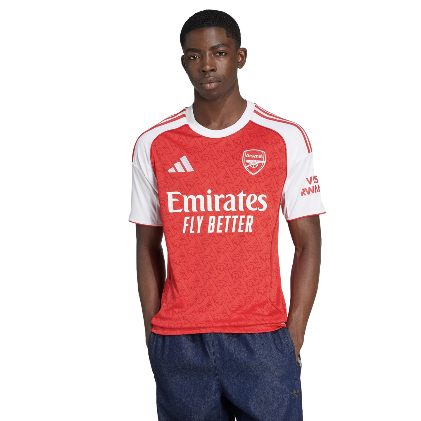 A man models the Arsenal home jersey, featuring a red body with white sleeves and sponsors. He is wearing dark blue shorts against a plain white background.