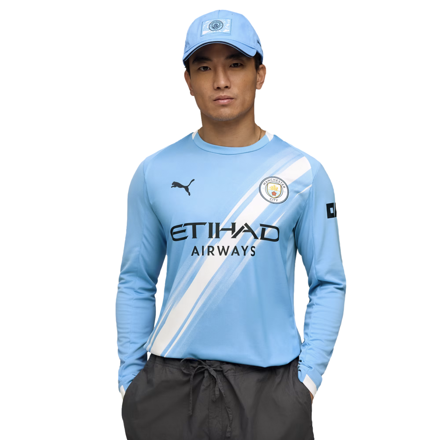 A man in a light blue Manchester City jersey and matching cap poses with his hands in his pockets against a white background.