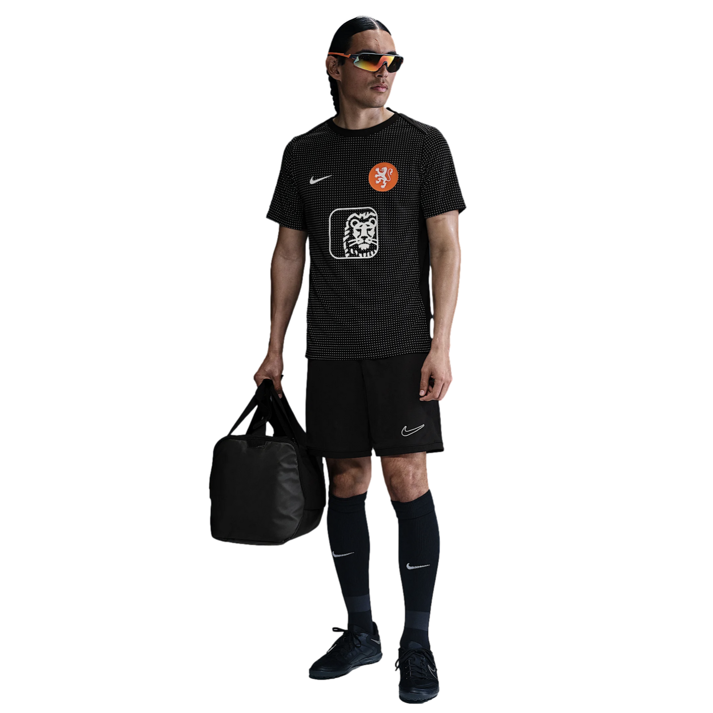 A man wearing black athletic clothing and sunglasses, holding a black duffel bag, stands against a plain white background.