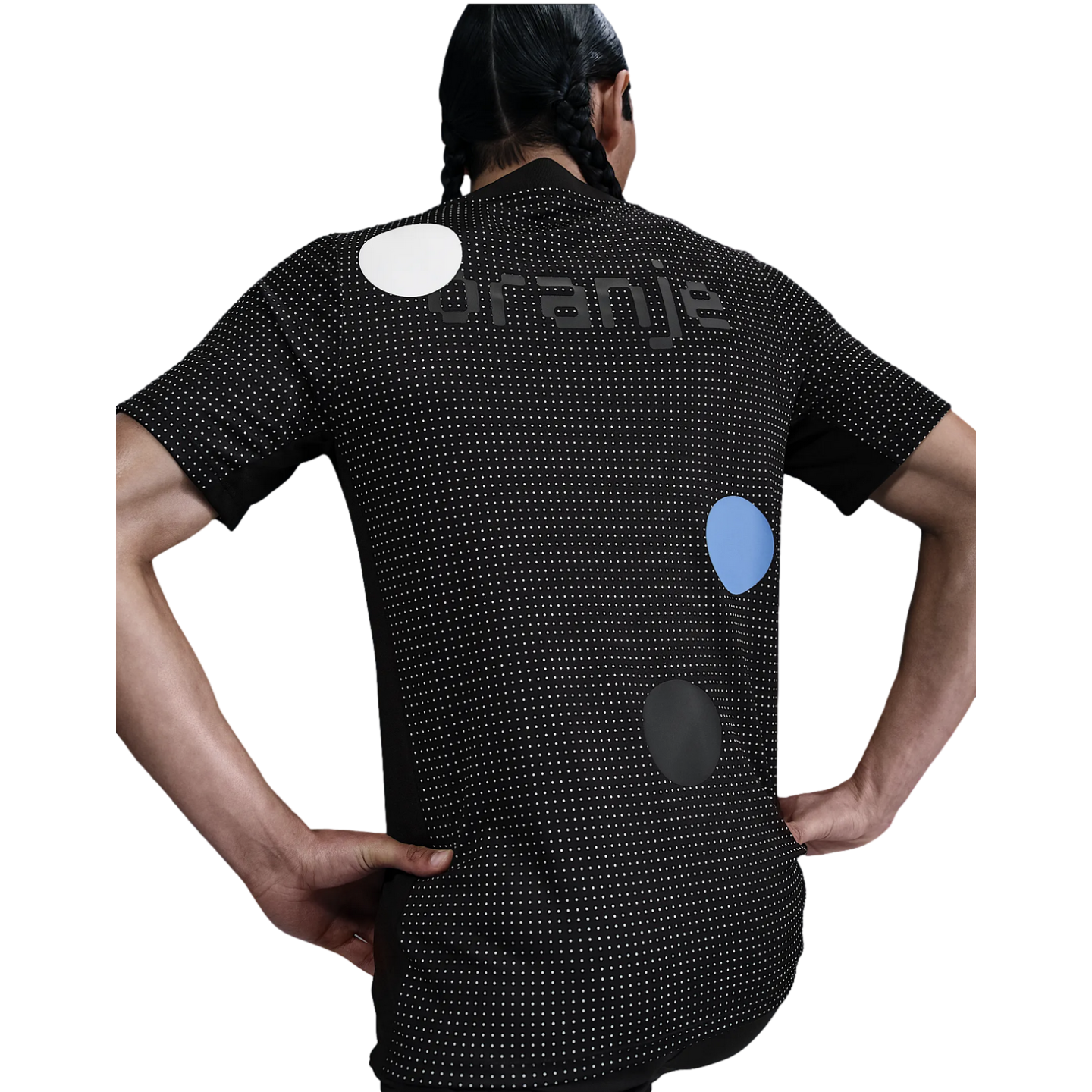 Rear view of a person with braided hair wearing a black jersey with white micro-dots, "ORANJE" text, and three large colored dots. Their hands are on their hips.