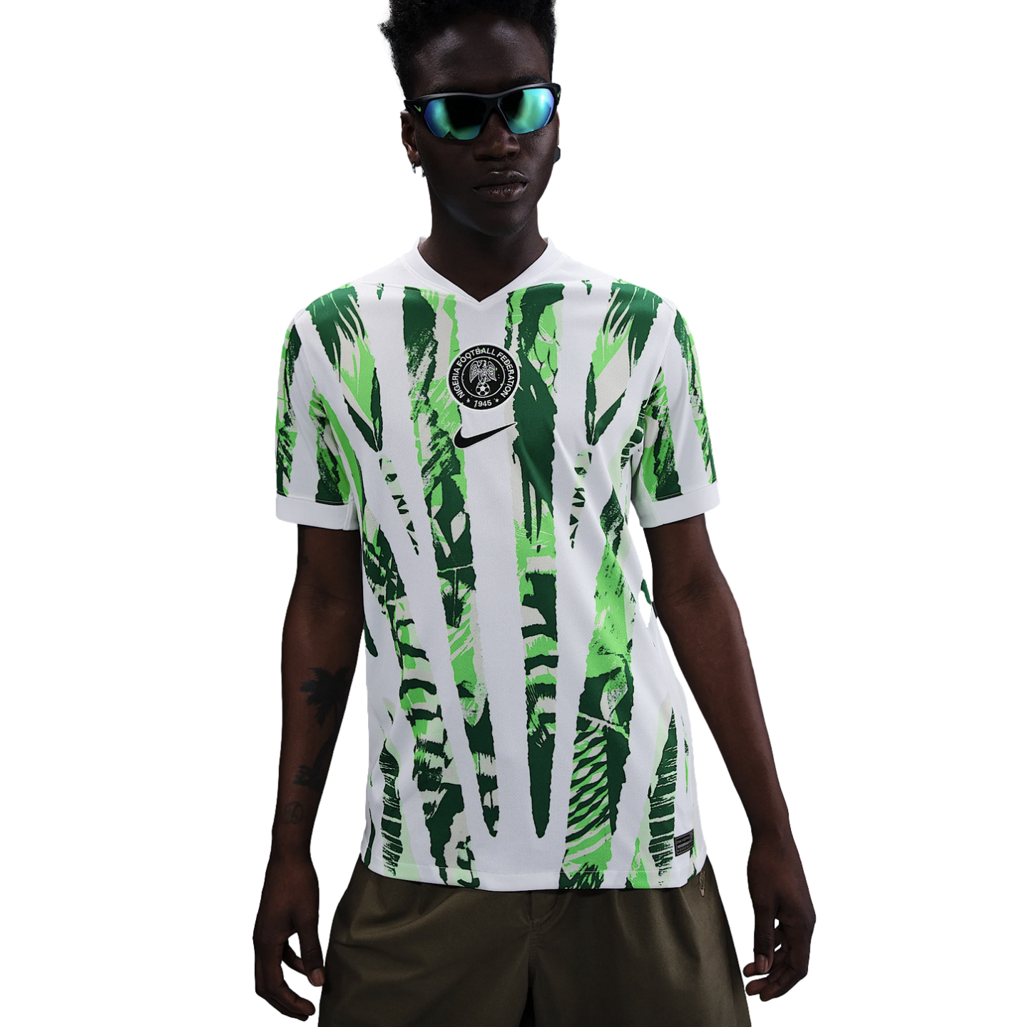 A man wears a patterned green and white Nike Nigeria football jersey and mirrored sunglasses against a white background.
