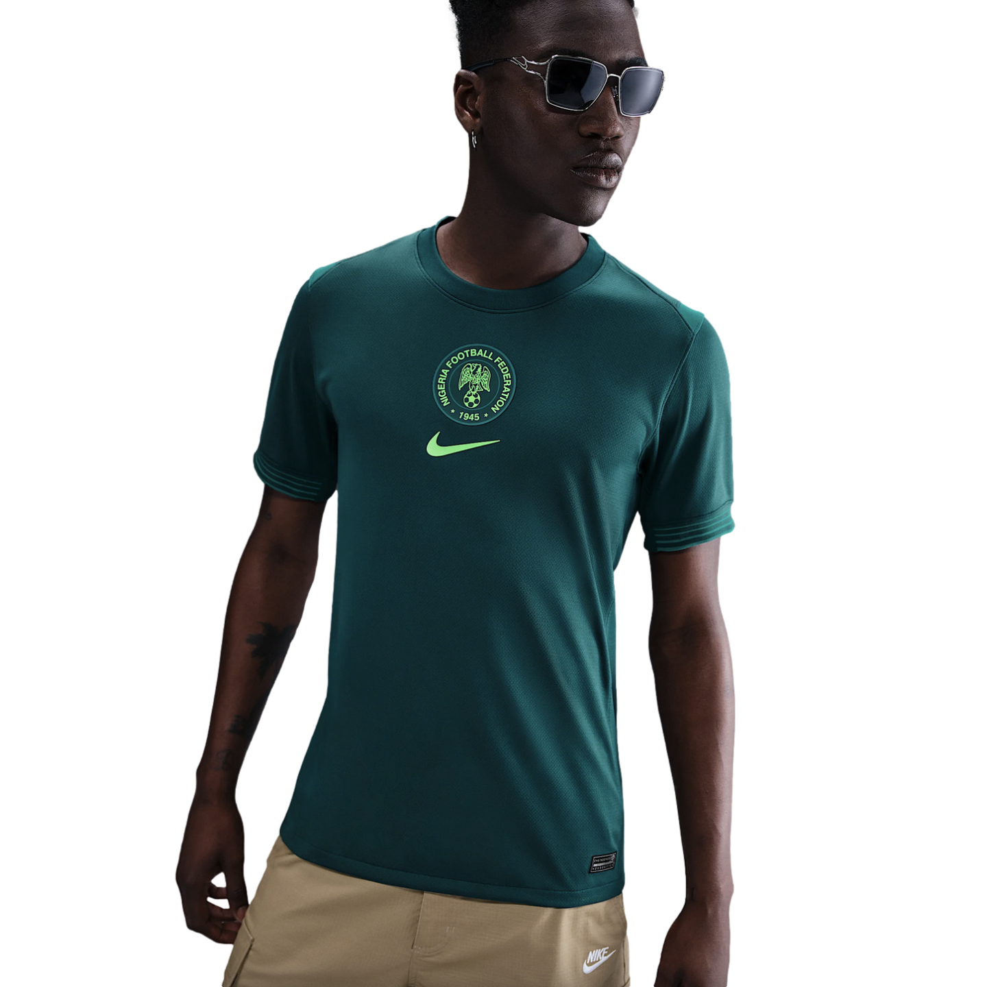 A young man models a dark green Nike Nigeria football shirt with a central crest and tan cargo pants against a white background.