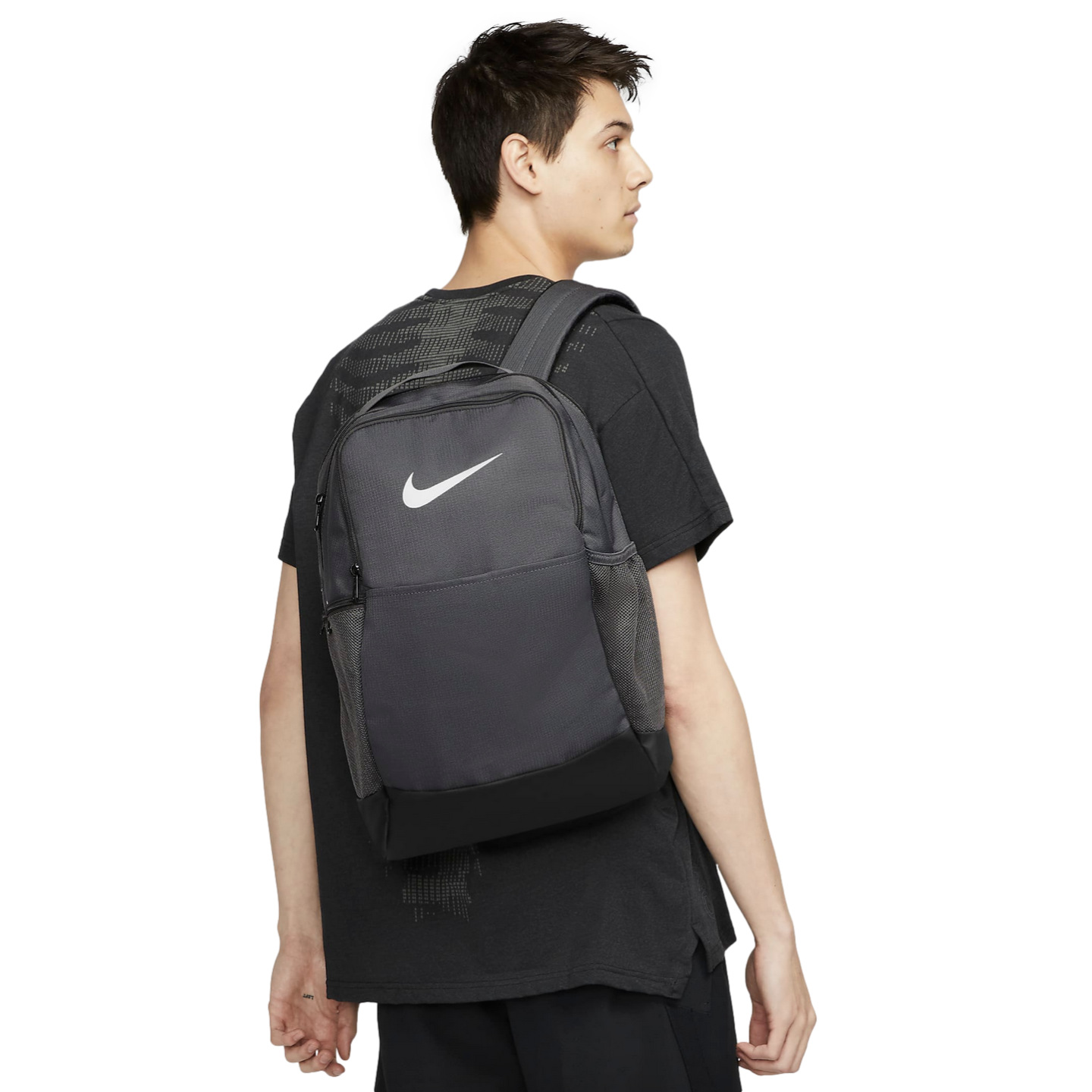 A man seen from behind wearing a black Nike t-shirt and carrying a dark grey Nike backpack over his right shoulder against a white background.