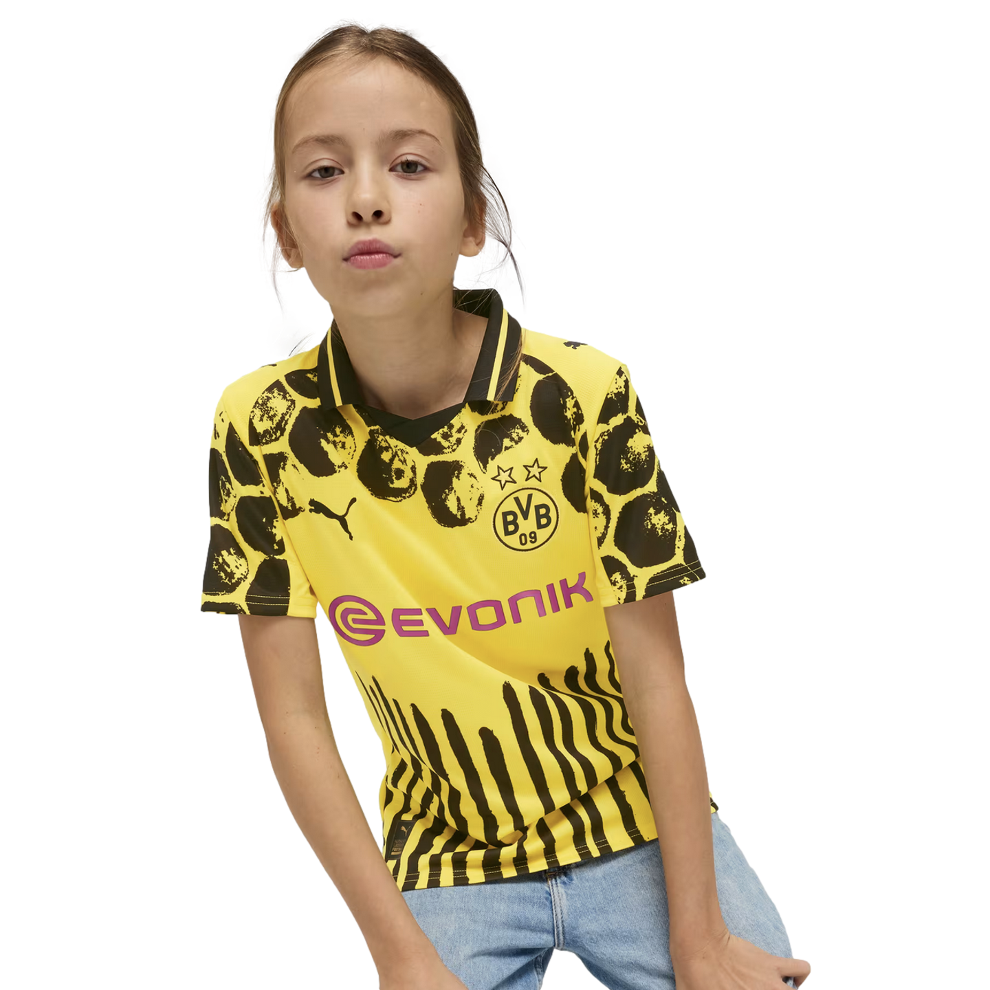 A young girl in a yellow and black Borussia Dortmund soccer jersey and blue jeans pouts against a plain white background.