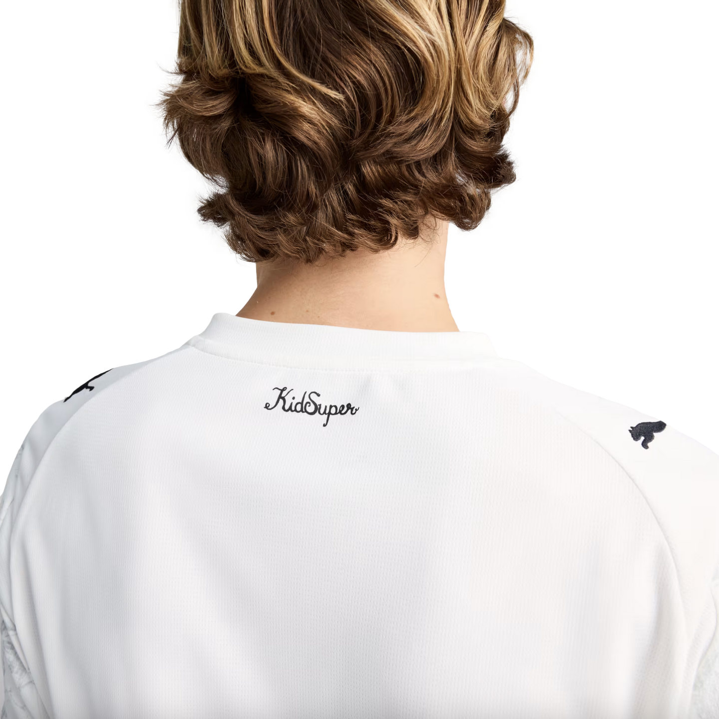 A rear view of a person with wavy brown hair wearing a white t-shirt featuring a cursive "KidSuper" logo on the upper back and Puma logos on the shoulders.