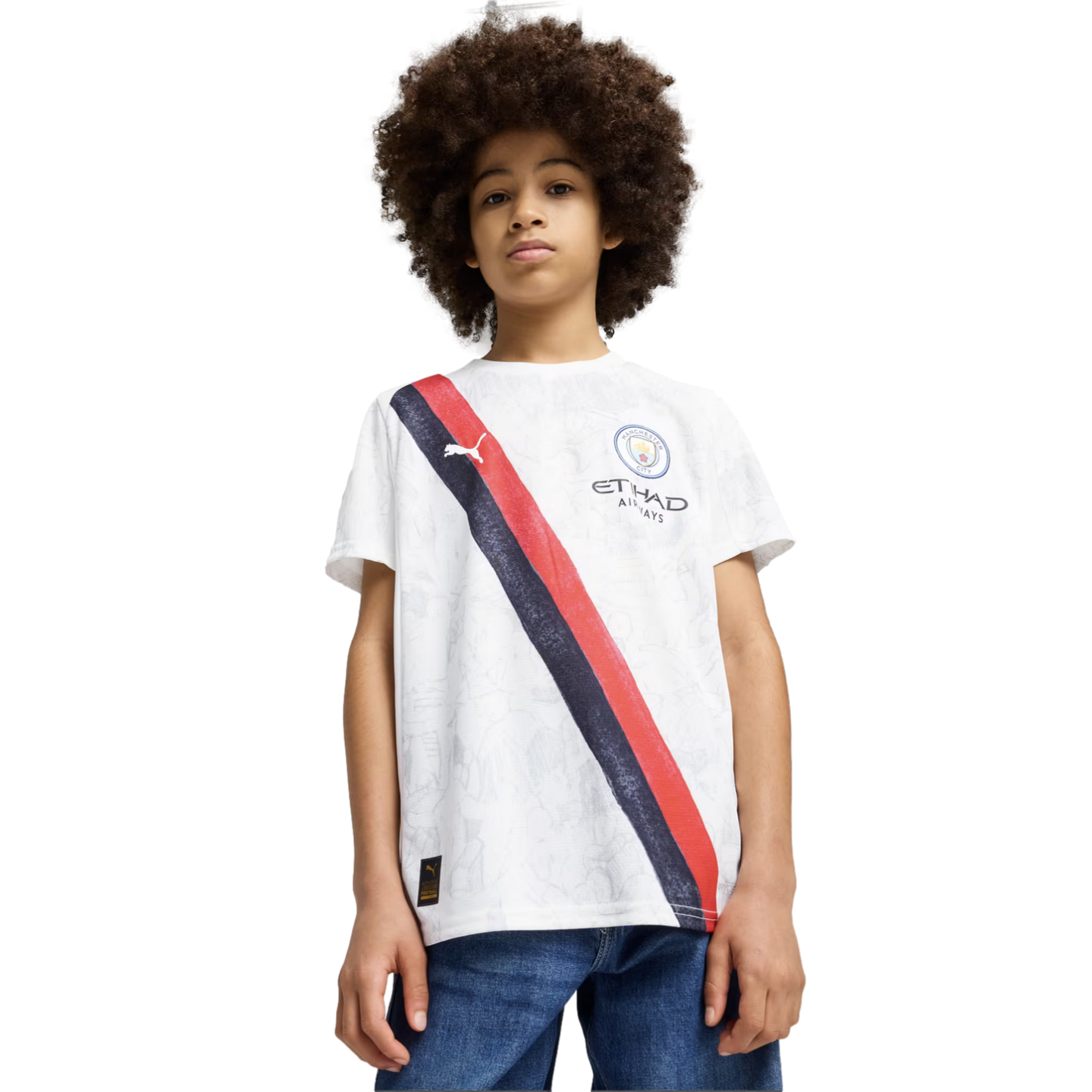 A boy with an afro wears a white Manchester City soccer jersey with a red and navy diagonal stripe, paired with blue jeans, against a plain white background.
