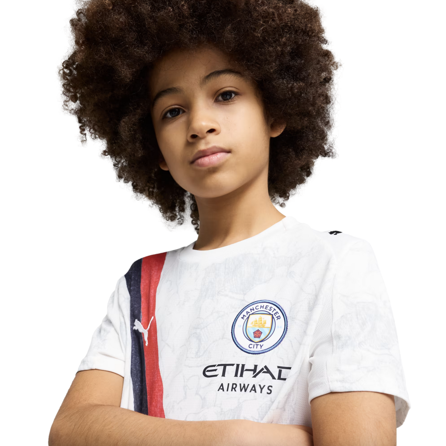 A young child with a large afro wears a white Manchester City soccer jersey, looking at the camera with arms crossed against a plain white background.