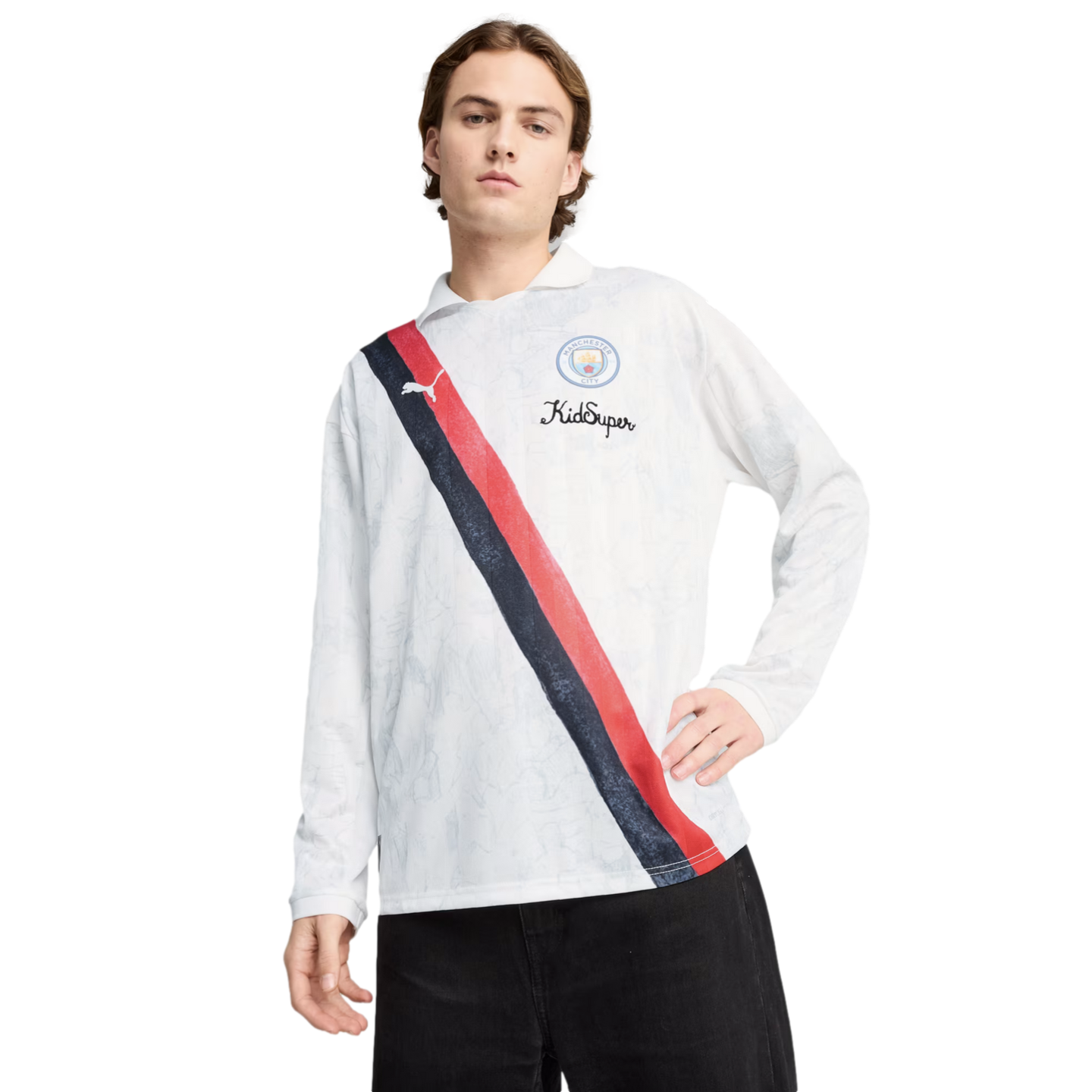 A man in a white Puma x KidSuper Manchester City long-sleeve polo featuring a red and navy sash, paired with black pants.