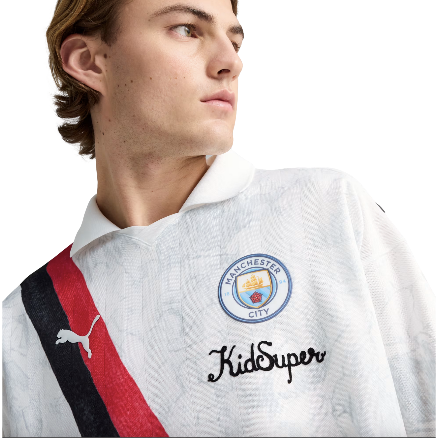 A young man wearing a white Manchester City x KidSuper Puma jersey featuring a red and black diagonal sash and the club crest.