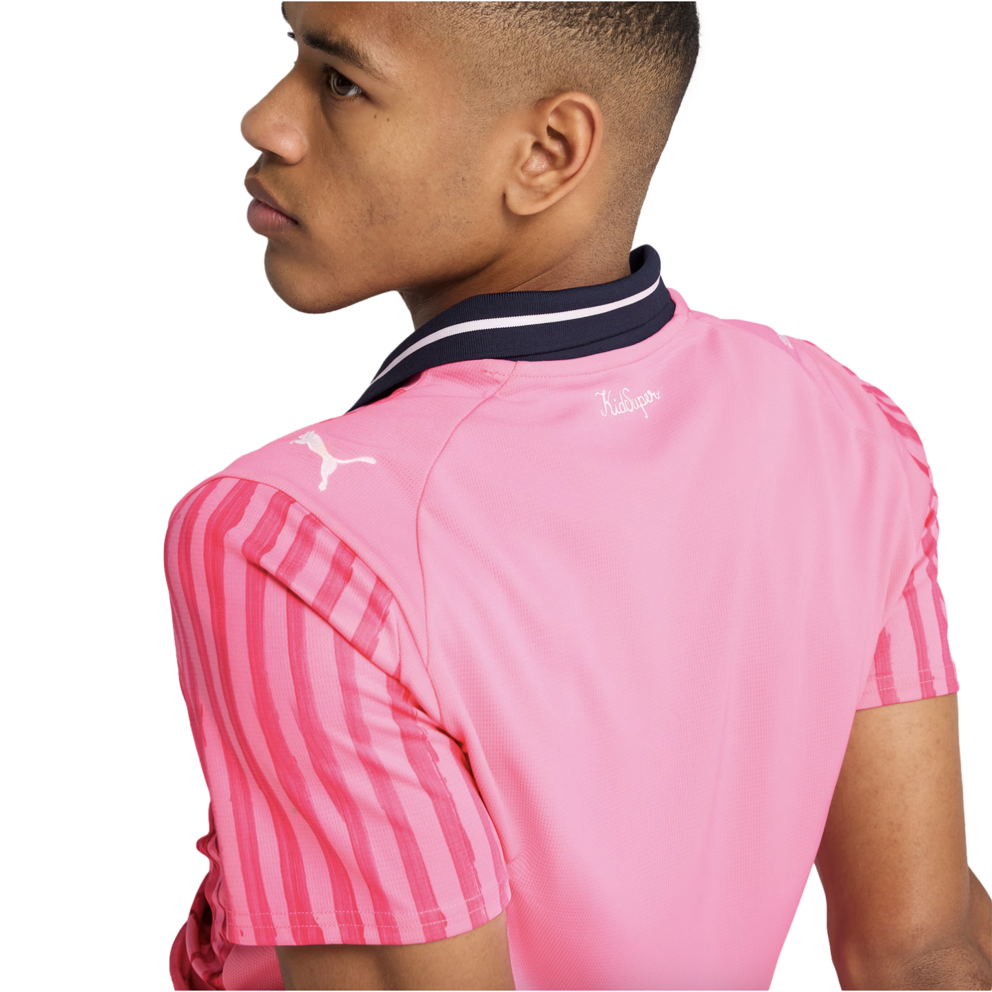 Side profile of a person wearing a pink Puma jersey with red striped sleeves and a navy blue collar. The jersey features a white Puma logo and "KidSuper" text on the back.