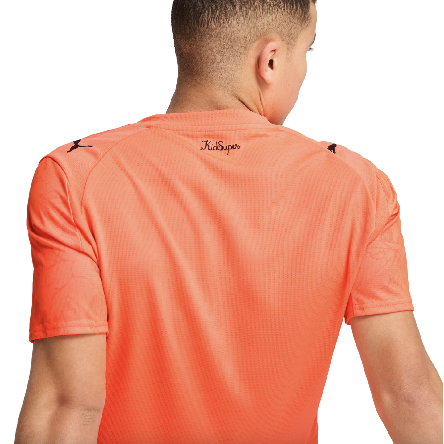 Back view of a person wearing an orange Puma x KidSuper t-shirt with logos on the shoulders and branding on the back collar.
