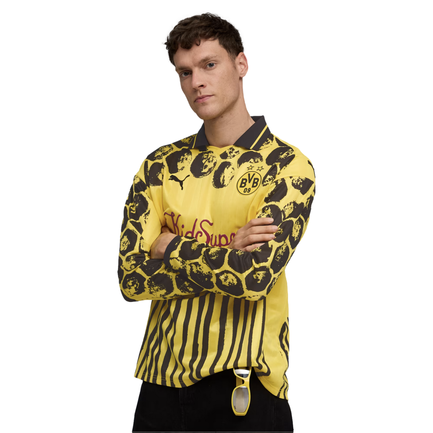 A man models a long-sleeved yellow and black PUMA x BVB x KidSuper jersey with varied patterns, including stripes and circles, standing with arms crossed against a white background.
