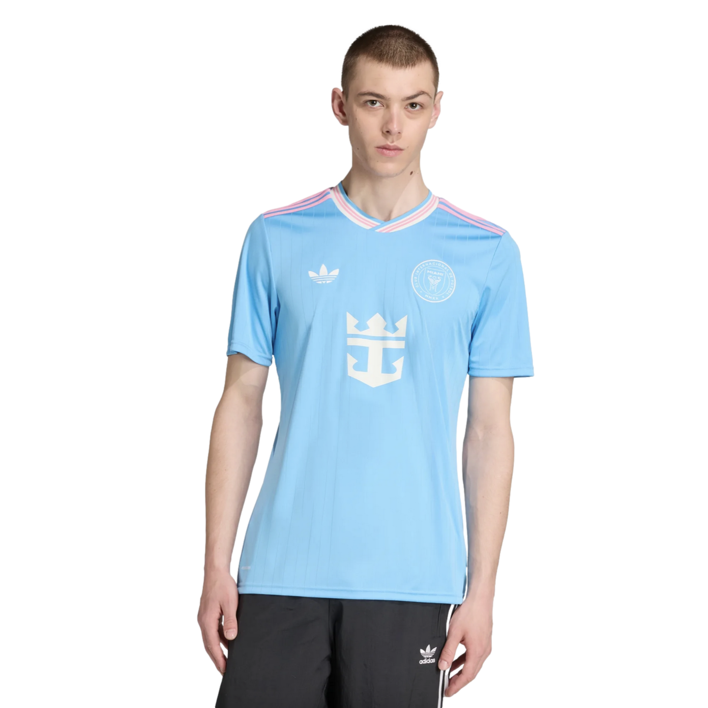 A man models a light blue Inter Miami CF jersey with pink accents and a central Royal Caribbean logo, paired with black Adidas shorts, on a white background.