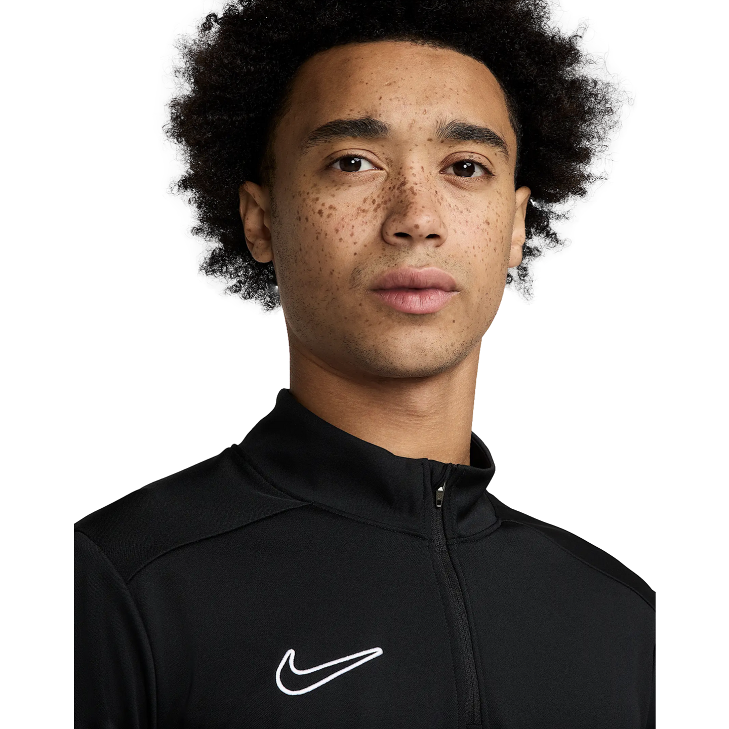 A close-up portrait of a young man with curly hair and freckles wearing a black Nike quarter-zip athletic top against a white background.
