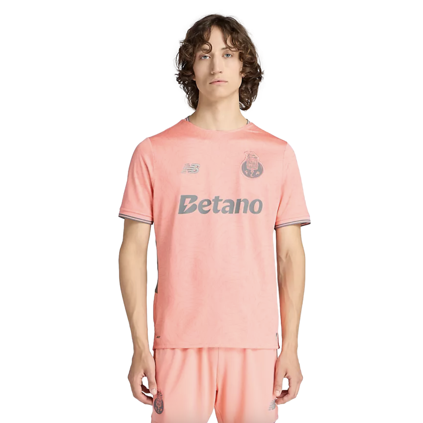 A man with wavy brown hair models a salmon-pink soccer kit with grey logos for "New Balance", "Betano", and a team crest on the chest.