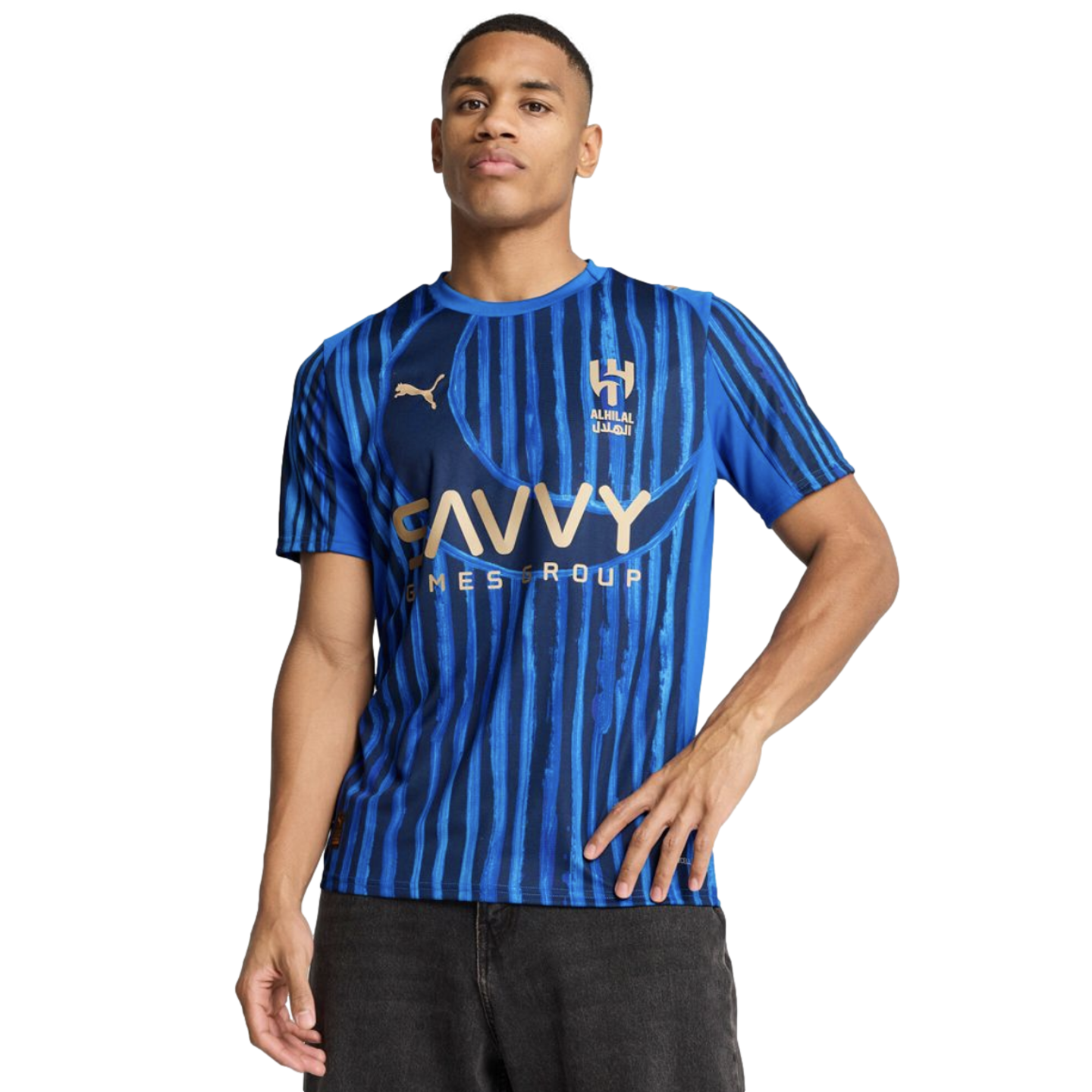 A man modeling an Al Hilal blue striped home soccer jersey with gold Puma and Savvy Games Group logos against a black background.