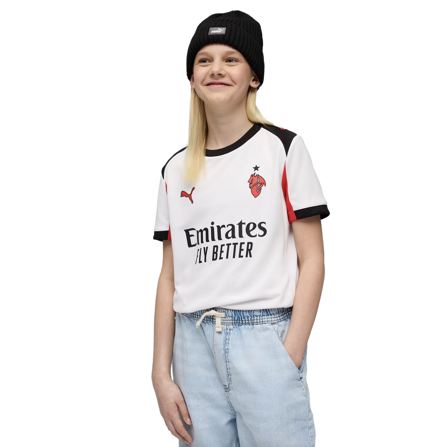 Smiling blonde youth in a black beanie, white AC Milan away jersey, and light blue jeans, standing against a black background.