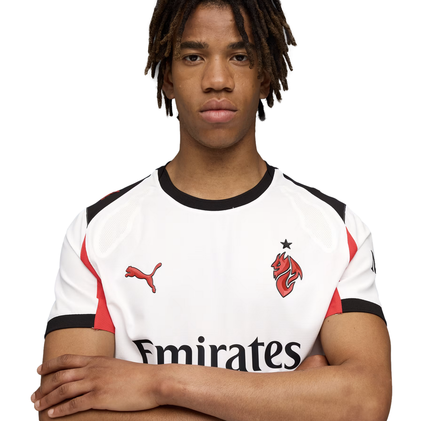 Portrait of a young man with dreadlocks wearing a white soccer jersey with red and black accents, posing with arms crossed against a solid black background.
