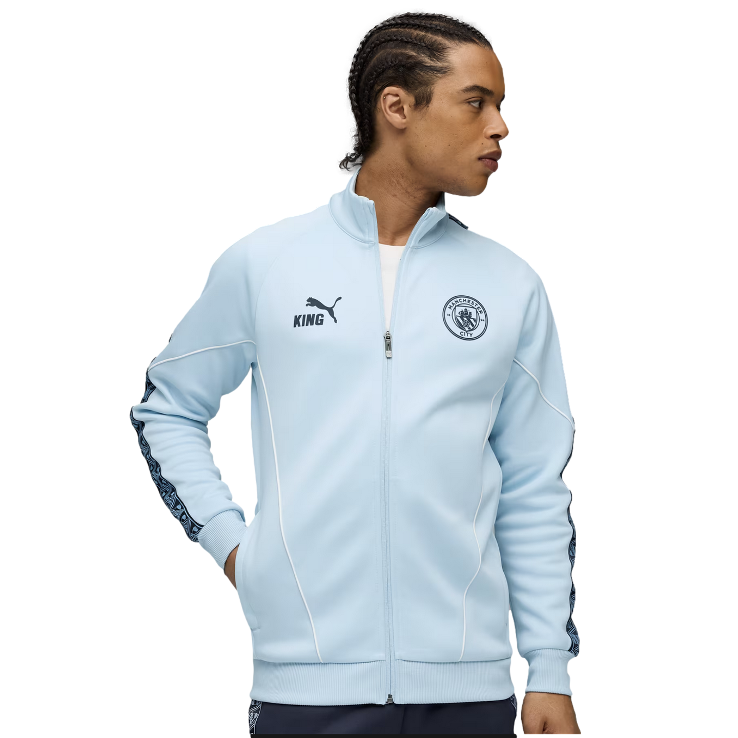 A man wearing a light blue Manchester City track jacket with the team crest, PUMA King logo, and patterned sleeve tape.