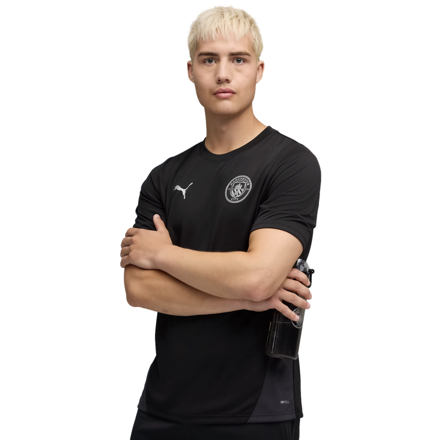 A man with blonde hair wears a black Puma Manchester City training shirt. He stands with arms crossed against a black background, holding a water bottle.