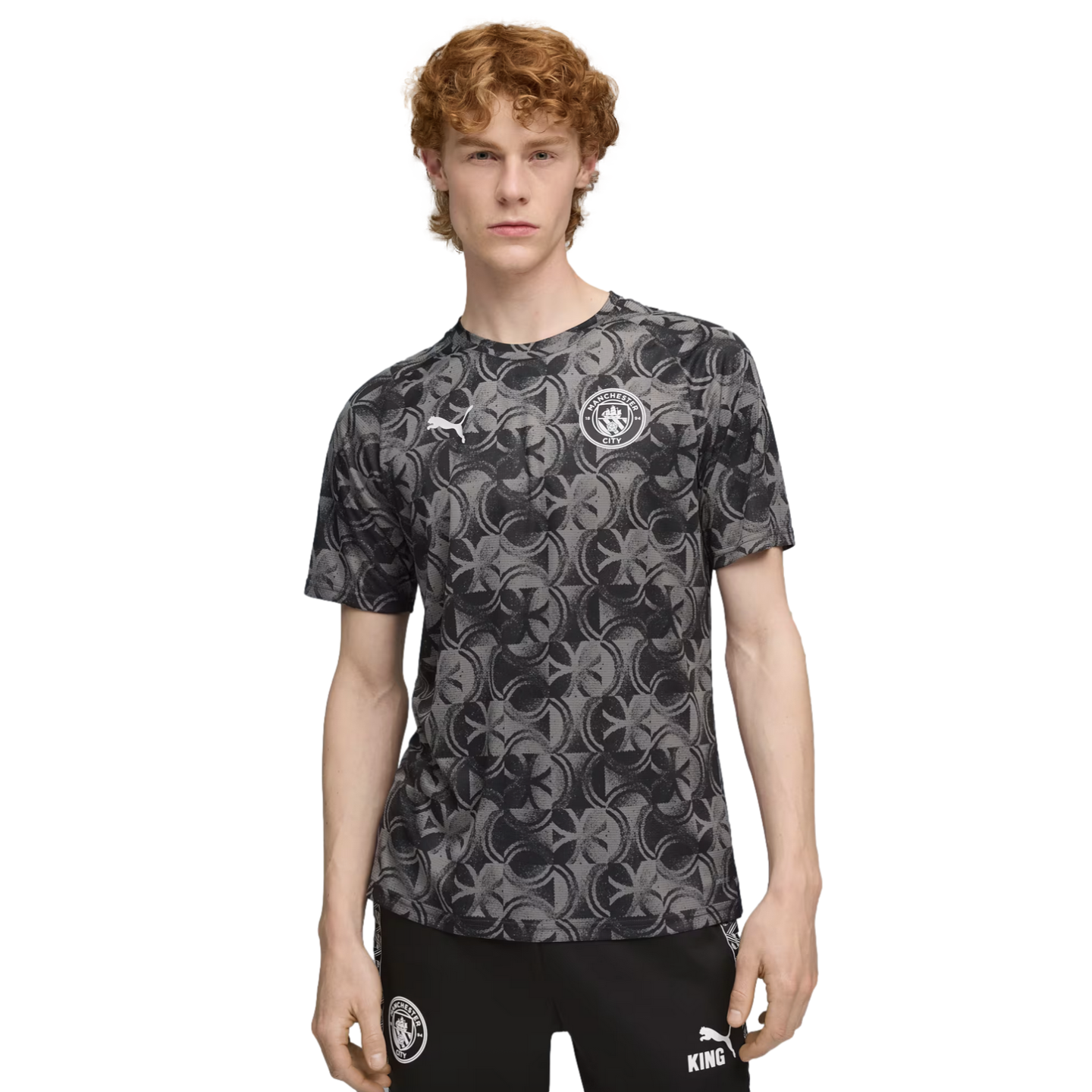 A young man with curly red hair wearing a grey patterned Manchester City jersey and black athletic pants against a black background.