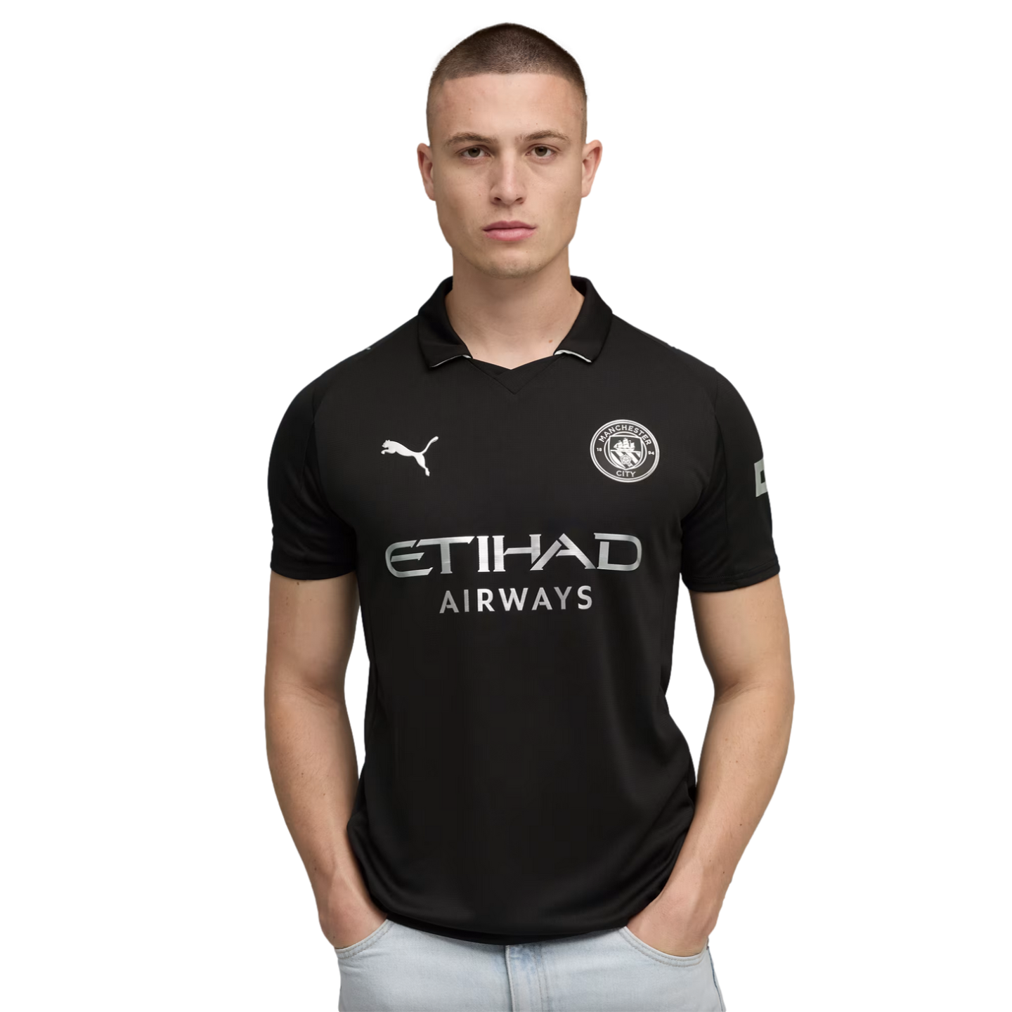 A man in a black Manchester City soccer jersey and light-wash jeans stands with his hands in his pockets against a solid black background.
