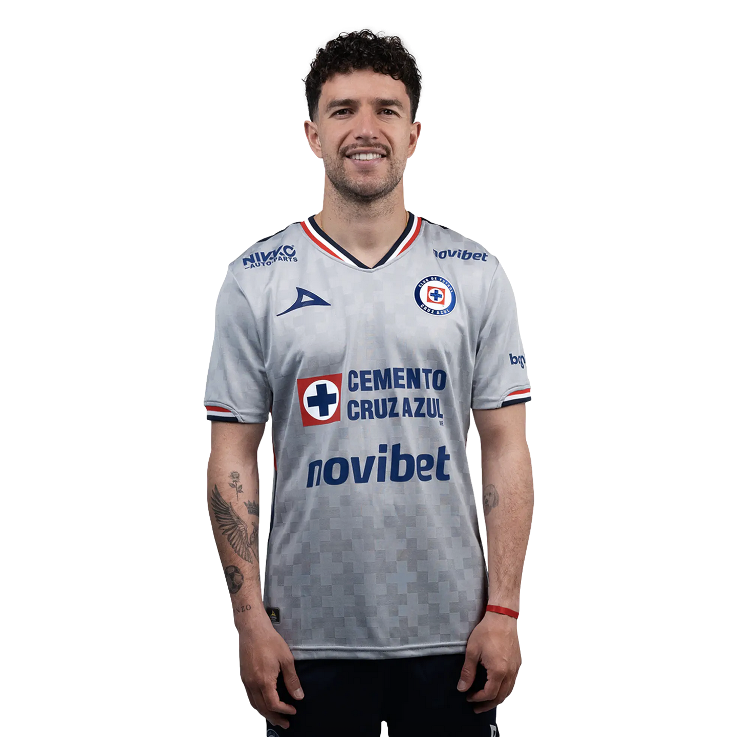 A smiling man with curly hair wearing a grey Cruz Azul soccer jersey with sponsor logos, against a black background. He has visible tattoos on his right arm.