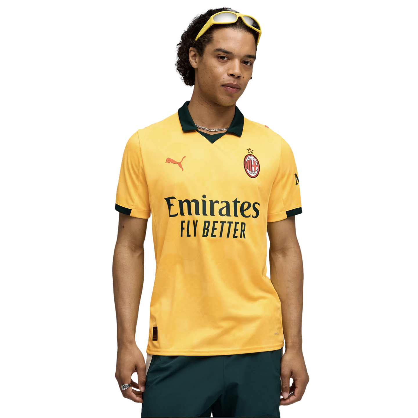 A man wearing a yellow AC Milan soccer jersey, dark green shorts, and yellow sunglasses on his head, posed against a black background.