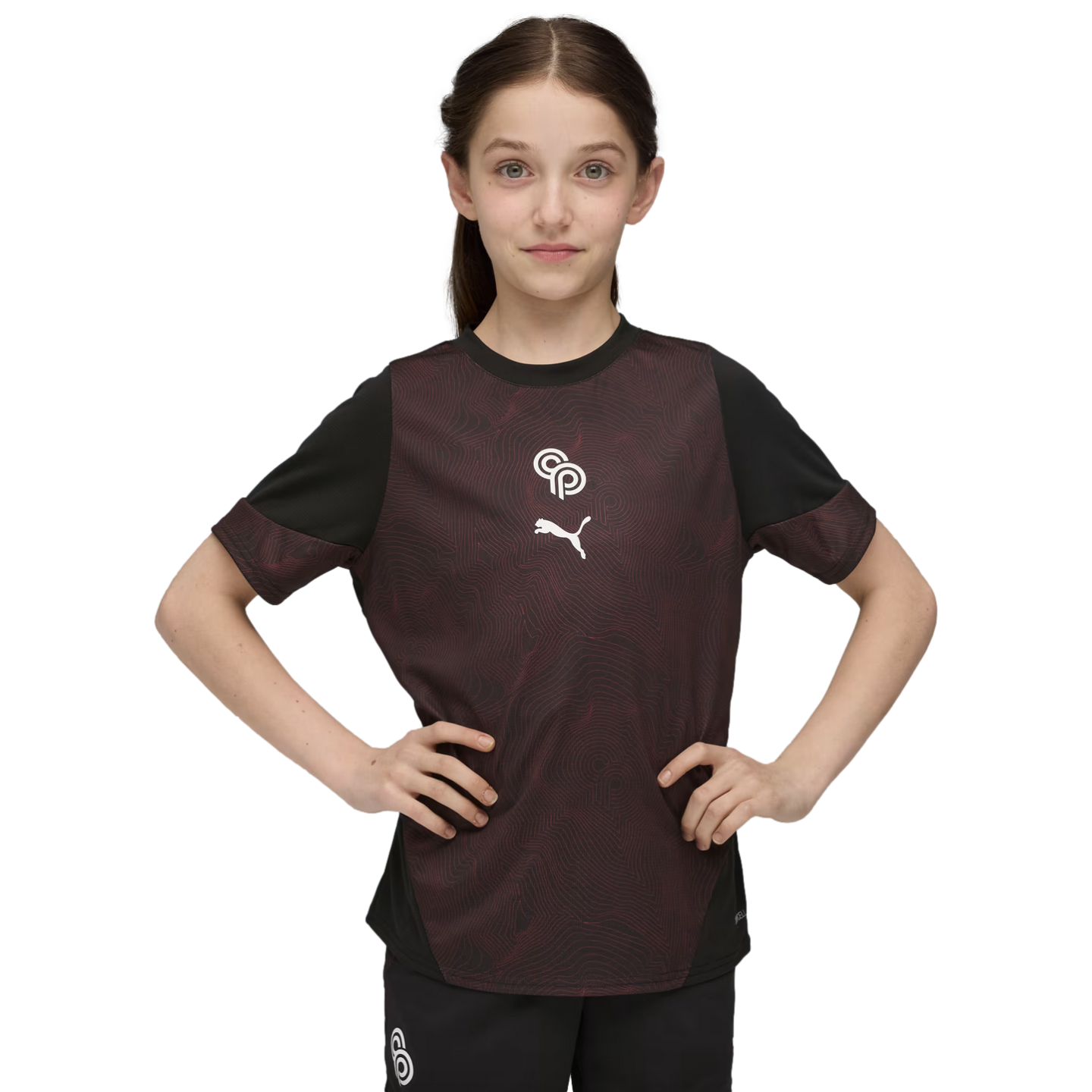 A young girl stands with hands on her hips wearing a maroon and black Puma athletic shirt against a solid black background.