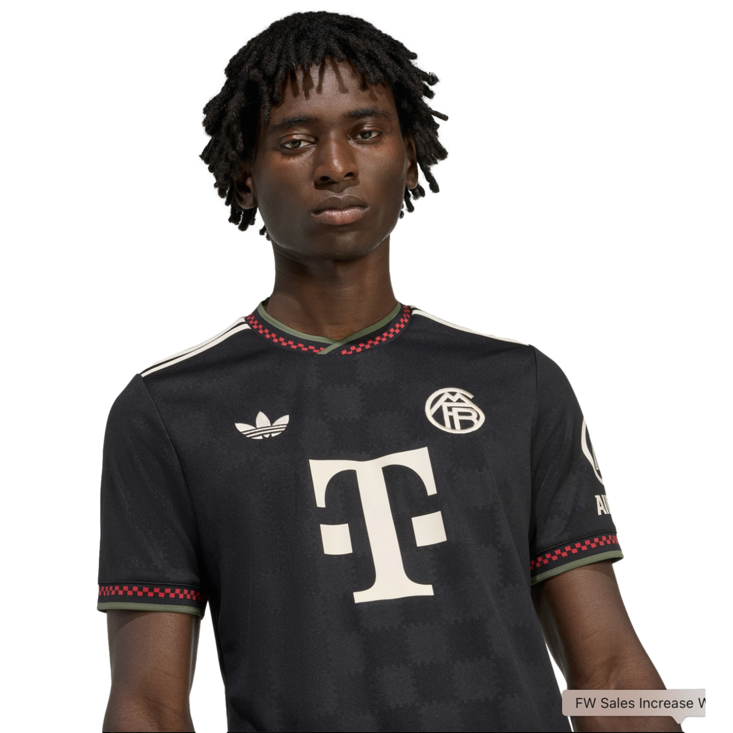 A Black man with dreadlocks wears a black FC Bayern Munich football jersey featuring a large white "T" logo and red checkered trim.