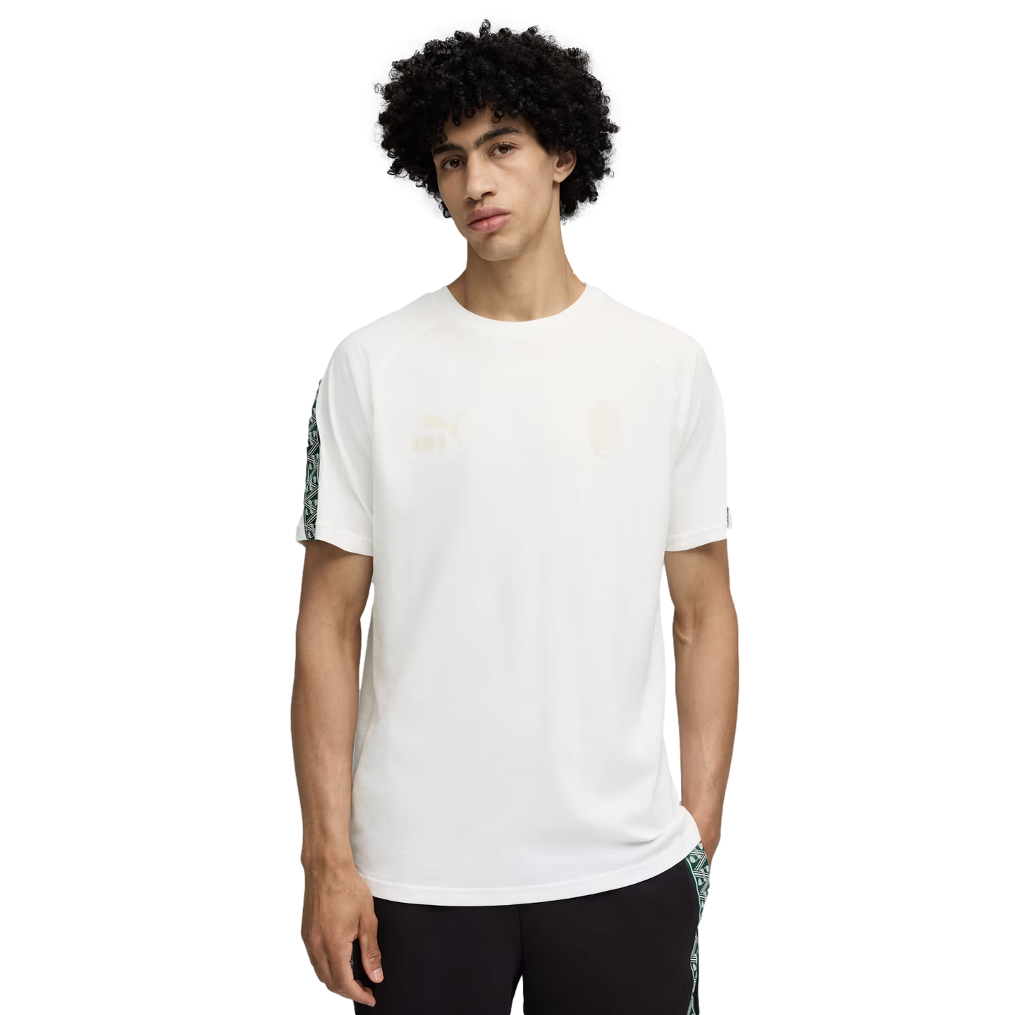 A curly-haired man stands against a black background wearing a white Puma short-sleeved t-shirt and dark trousers with patterned side details.