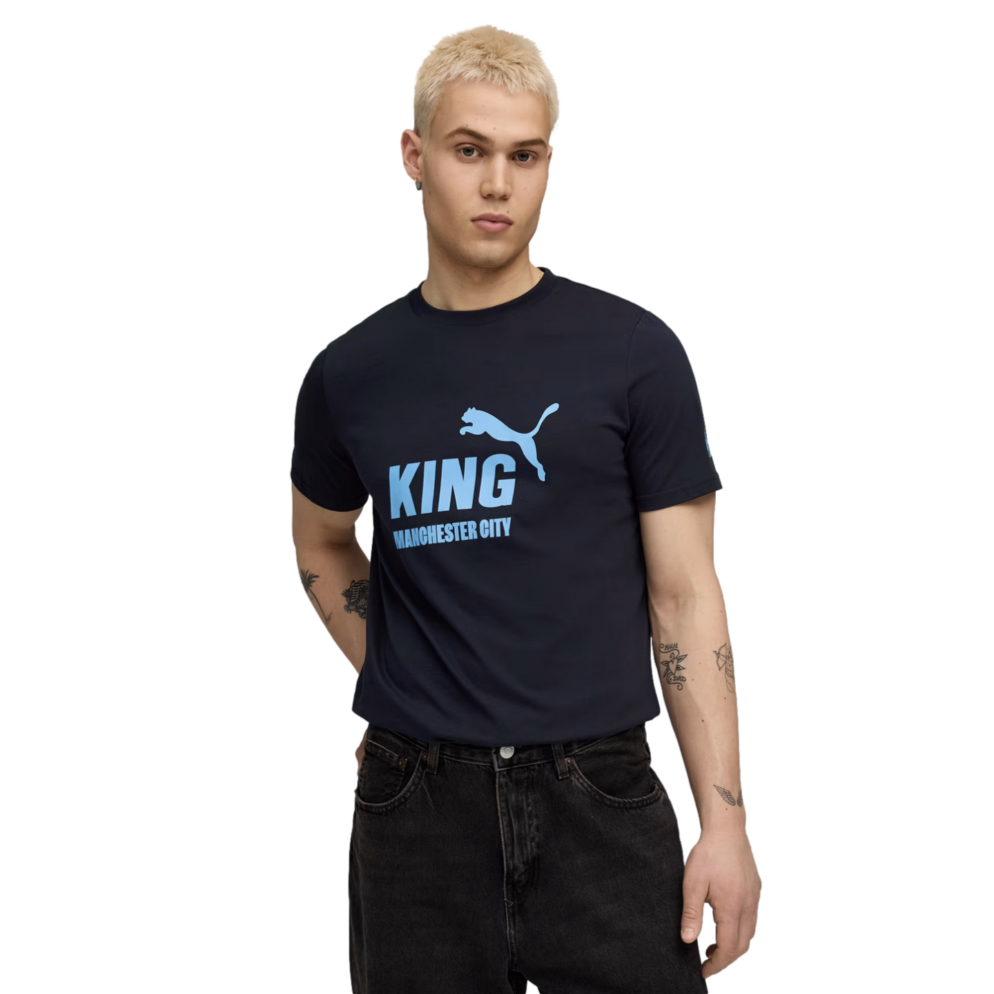 A person with short blonde hair wears a dark blue Manchester City T-shirt and black jeans against a black background.