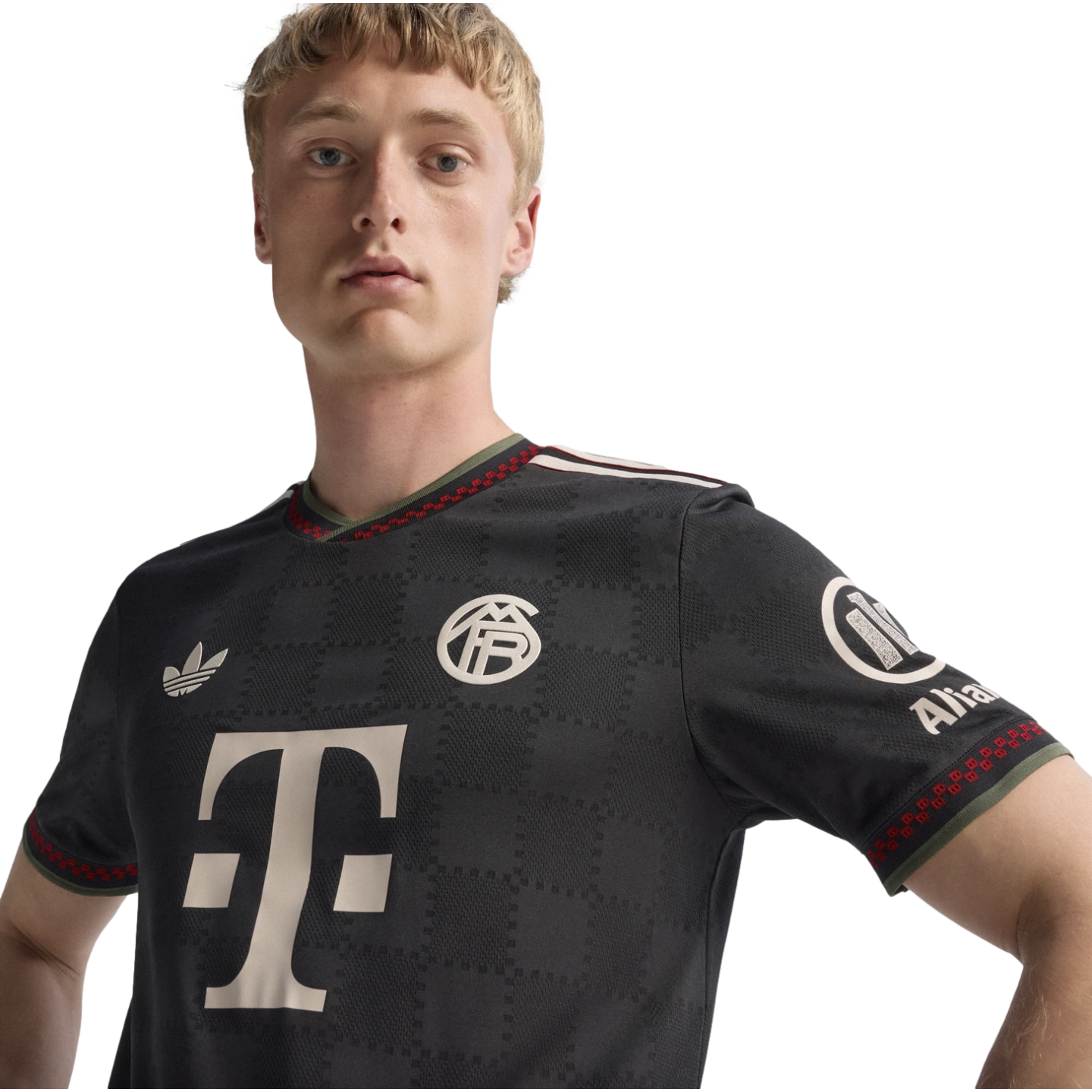 A blonde man wearing a dark gray Bayern Munich soccer jersey with a large white "T" logo and a retro club crest against a black background.