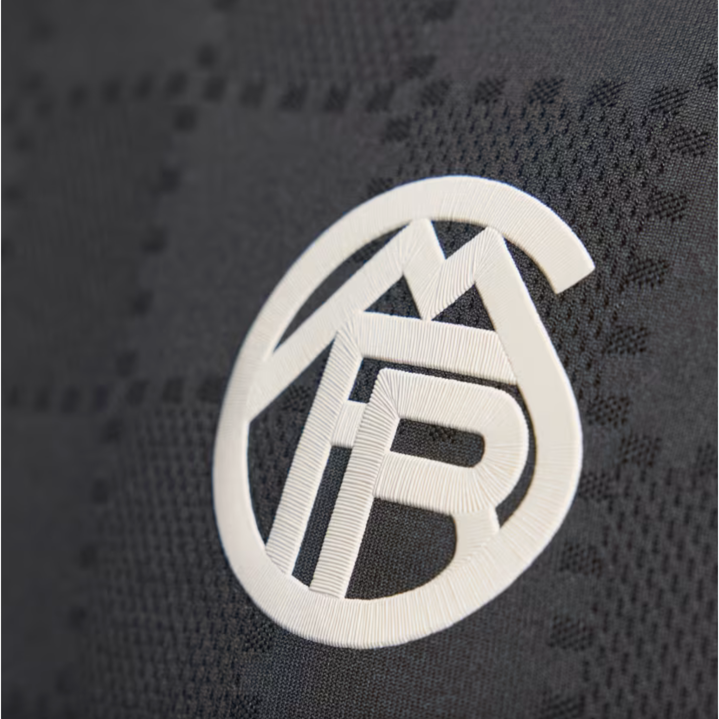 White circular monogram logo with a raised texture on dark grey fabric featuring a subtle geometric pattern.